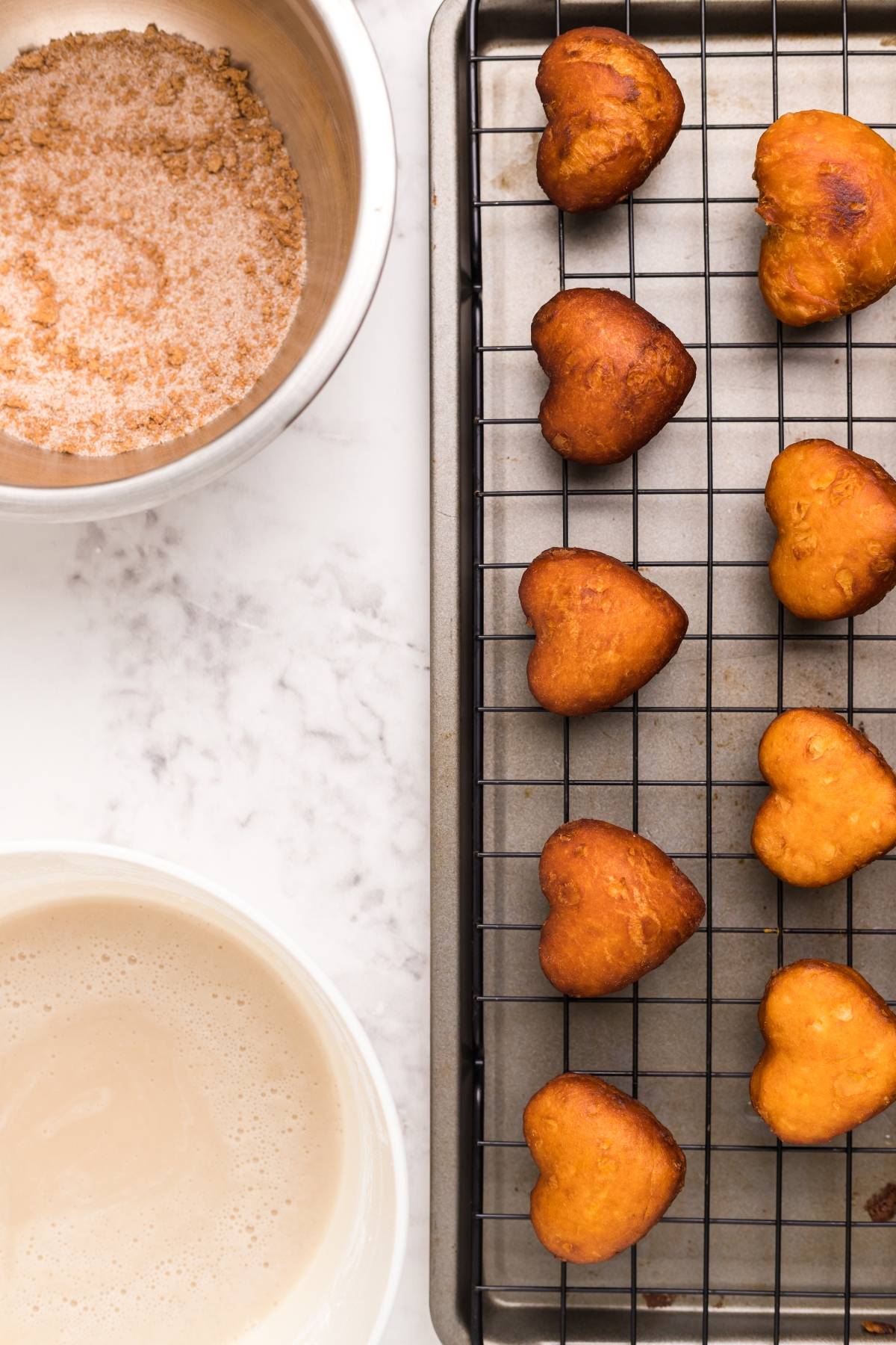 Heart-shaped donuts on a cooling rack, with bowls of cinnamon sugar and glaze on a marble countertop—perfect for trying your favorite mini doughnut recipe or testing out a new cinnamon roll hack.