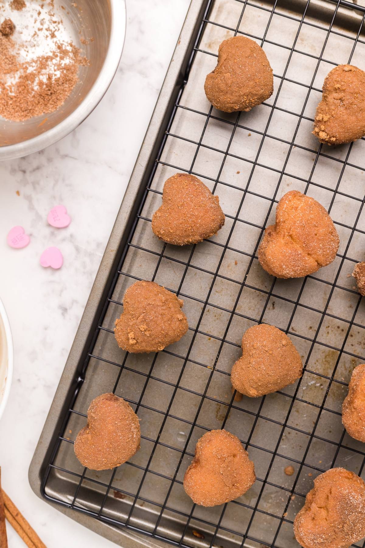 Heart-shaped cookies on a cooling rack, with bowls of ingredients and pink heart sprinkles nearby—perfect inspiration for your next heart shaped doughnut holes or mini doughnut recipe.