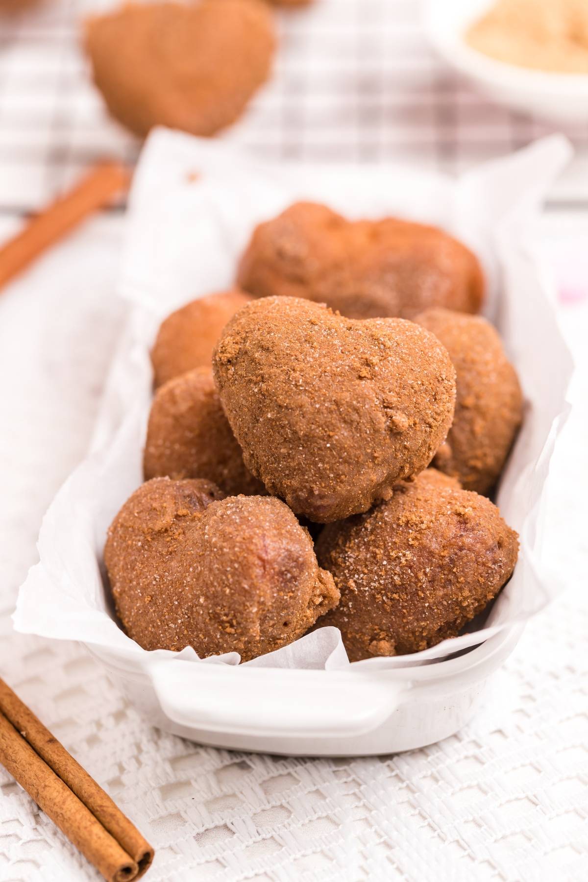 Heart-shaped cinnamon sugar donuts in a white dish on a white surface, cinnamon sticks nearby—perfect for trying a mini doughnut recipe or your favorite cinnamon roll hack.