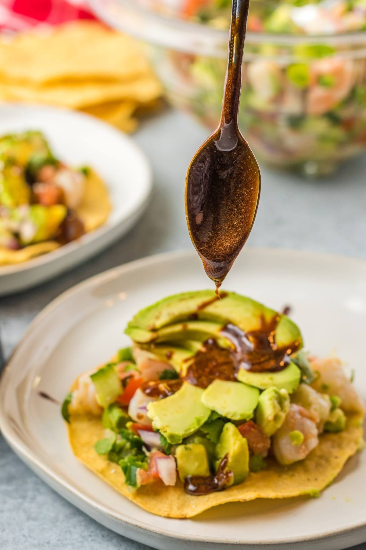 A spoon drizzles homemade unagi sauce over a tostada topped with avocado and ceviche on a white plate.