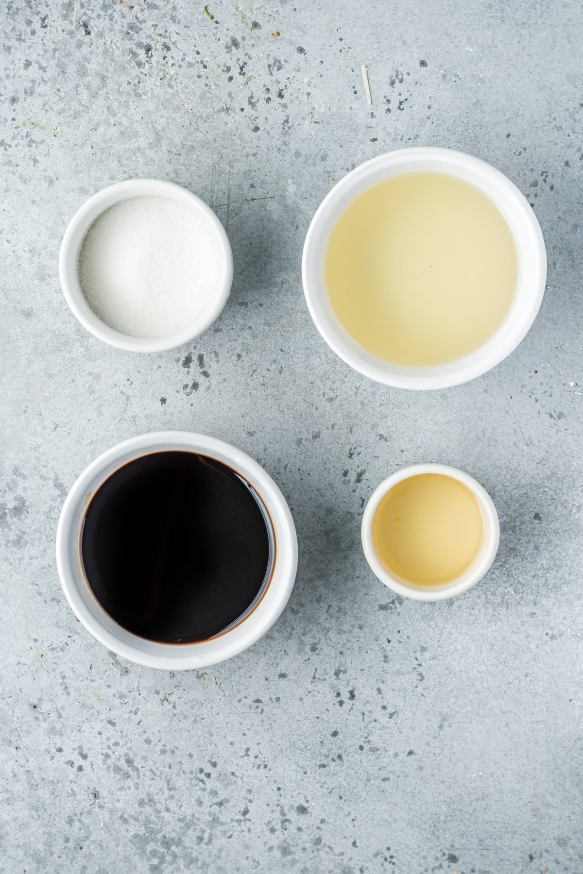 Four small white bowls containing sugar, soy sauce, rice vinegar, and mirin on a gray surface—perfect ingredients for homemade unagi sauce, a delicious choice as a sauce for seafood.
