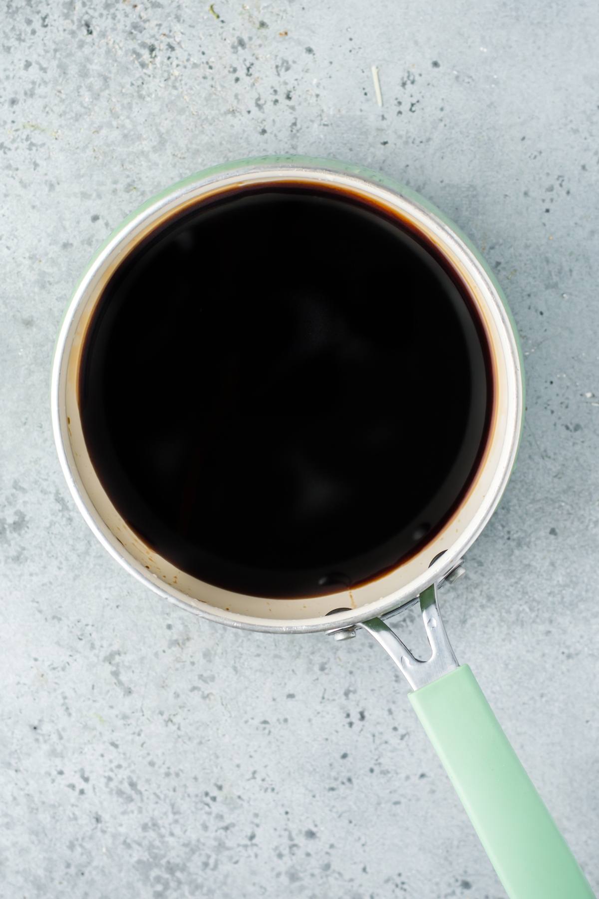 A saucepan filled with dark liquid, possibly homemade unagi sauce, sits on a light gray textured surface.