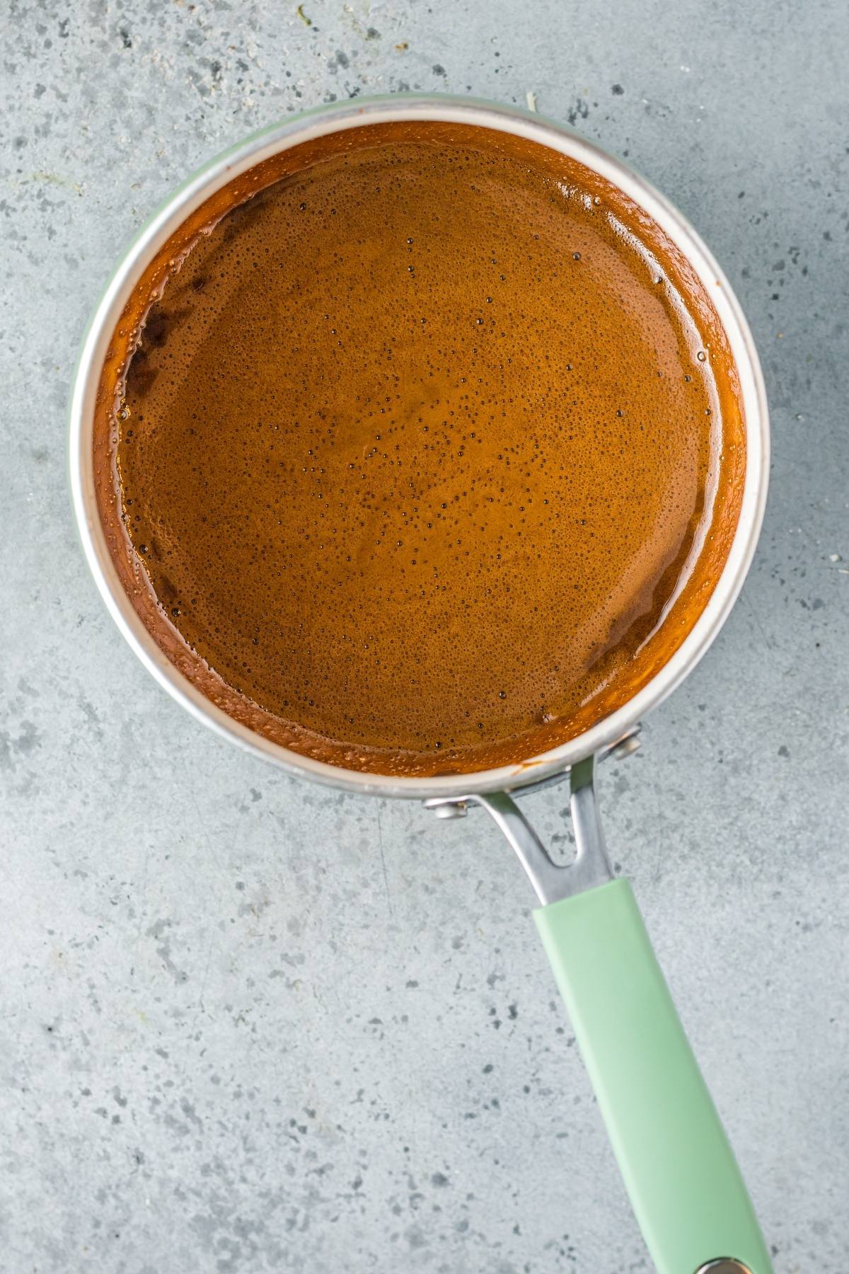 A saucepan with thick, frothy chocolate liquid on a light gray textured surface, perfect for pairing with Unagi sauce or as a unique twist alongside your favorite sauce for seafood.