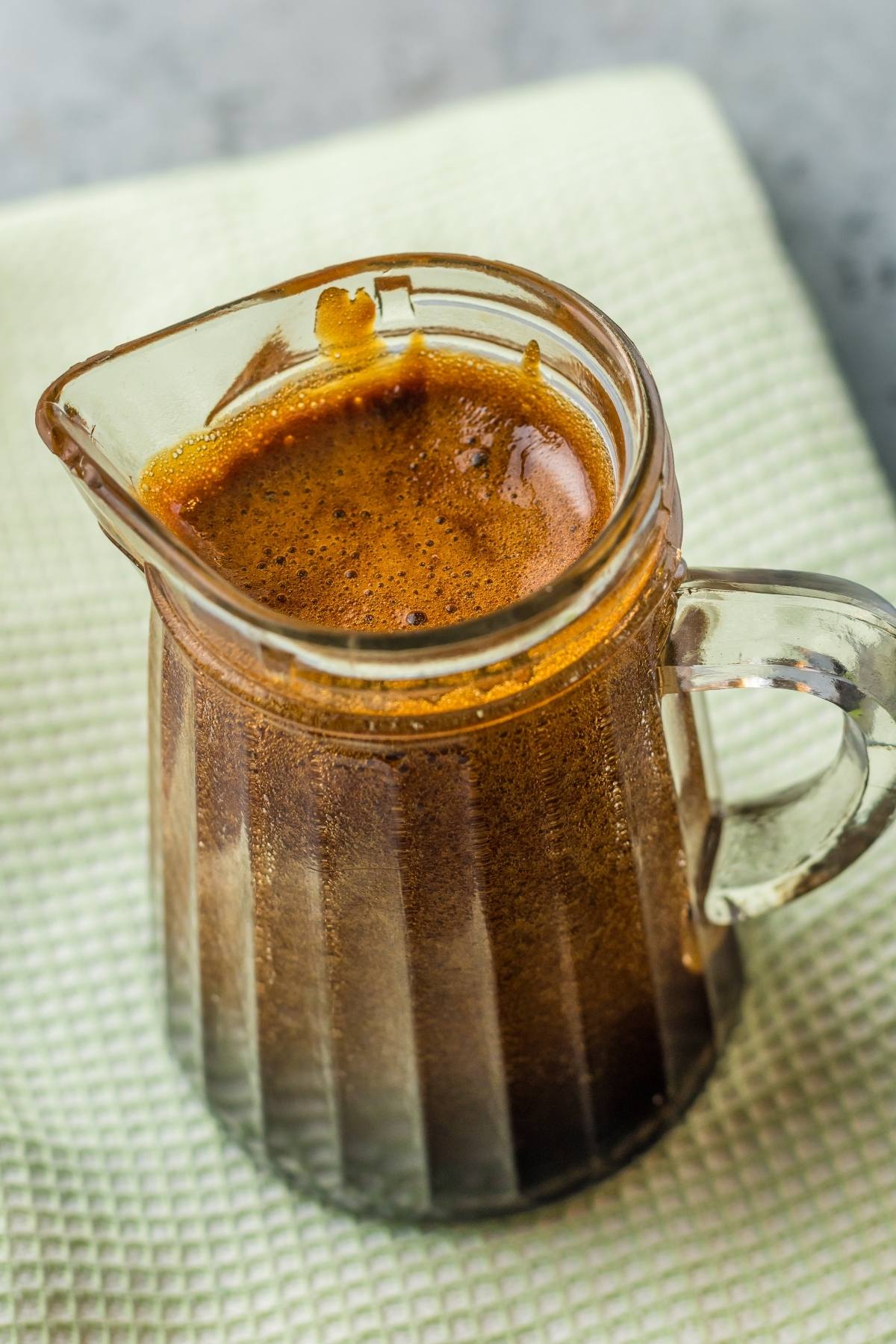 Small glass pitcher filled with dark brown espresso crema, resembling rich unagi sauce, sitting on a light textured surface.