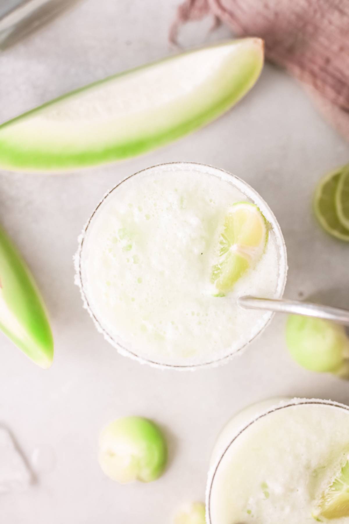 Overhead view of a frothy green honeydew margarita with a lime wedge in a glass, surrounded by fresh cucumbers and limes—an easy tequila cocktail perfect for summer sipping.