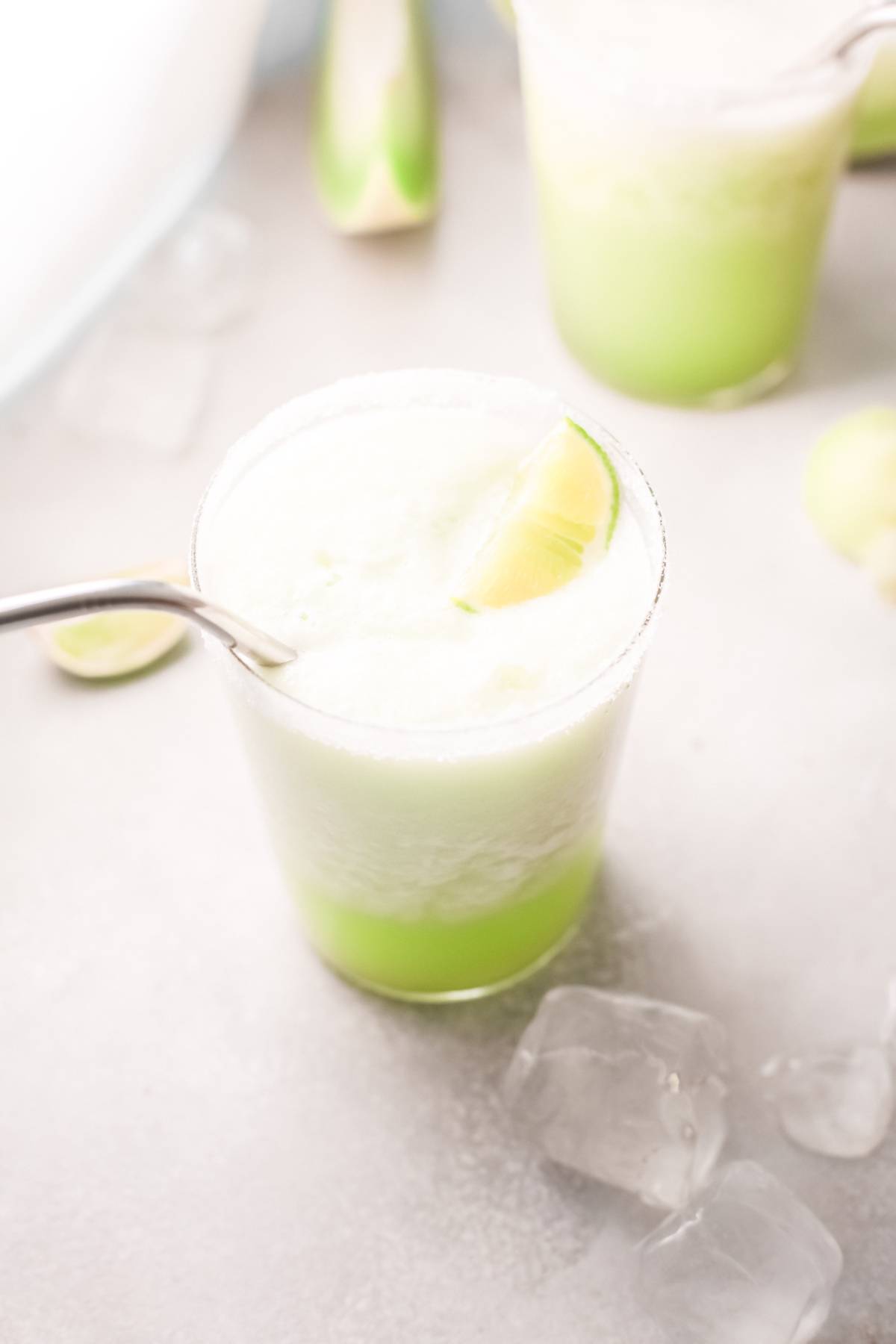 A glass of honeydew margarita with a frothy top, a lime wedge, and a metal straw sits surrounded by ice cubes—an easy tequila cocktail perfect for refreshing summer sips.