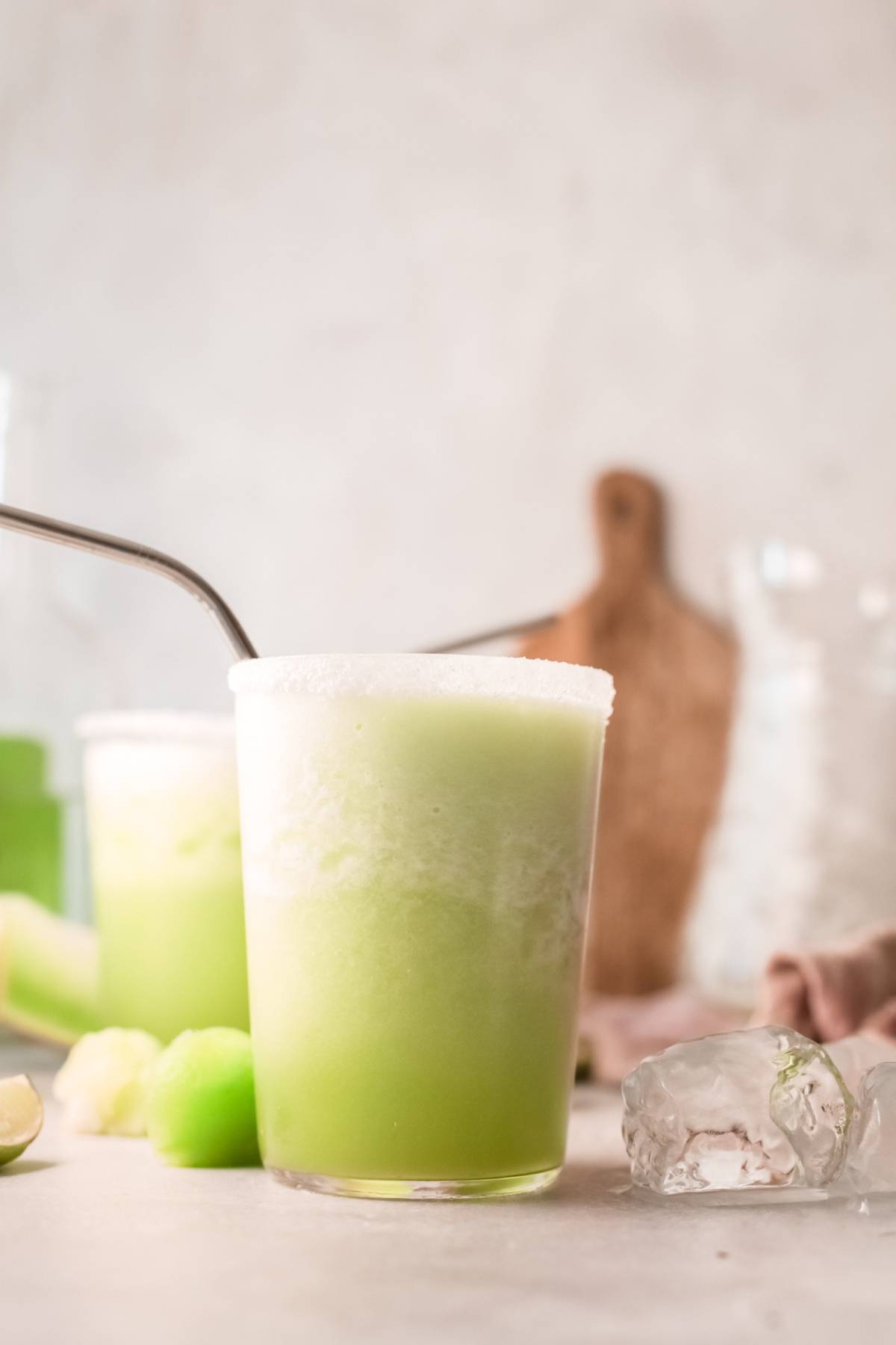A glass of green honeydew cocktail with a metal straw, ice, and melon pieces on a light surface—perfect for a refreshing summer cocktail recipe with tequila.
