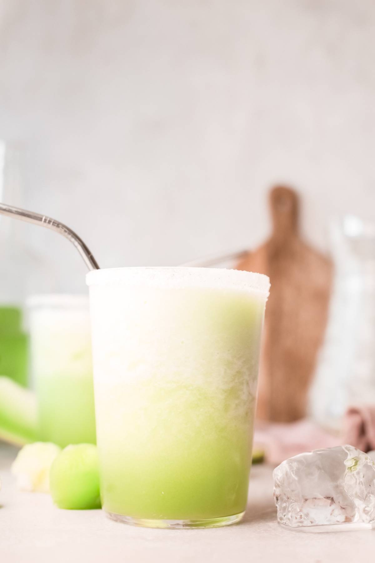 A glass of light green honeydew margarita smoothie with a metal straw, surrounded by ice cubes and melon pieces.