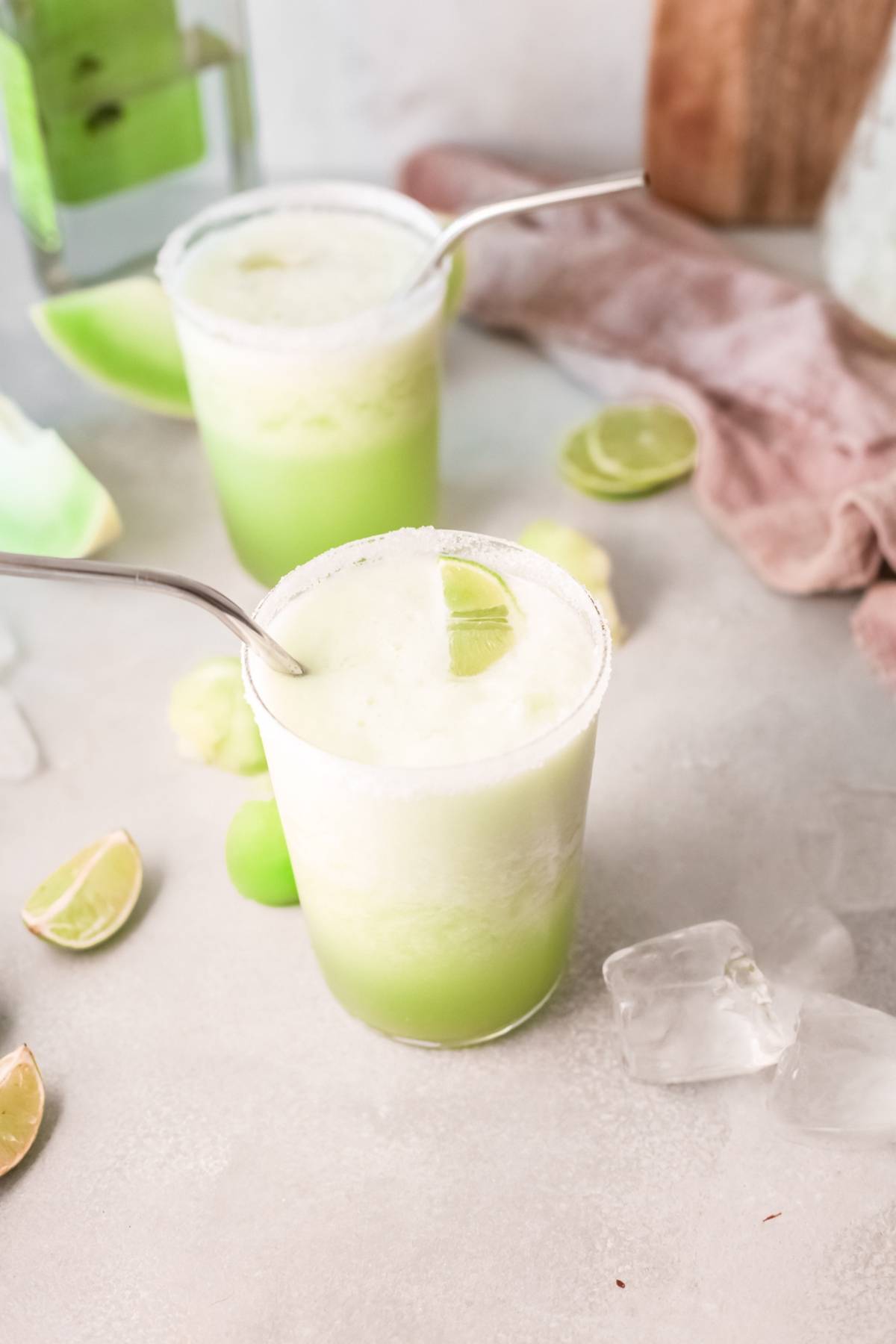 Two glasses of light green honeydew margarita with ice, lime wedges, and metal straws sit on a light surface—an inviting summer cocktail recipe with tequila.