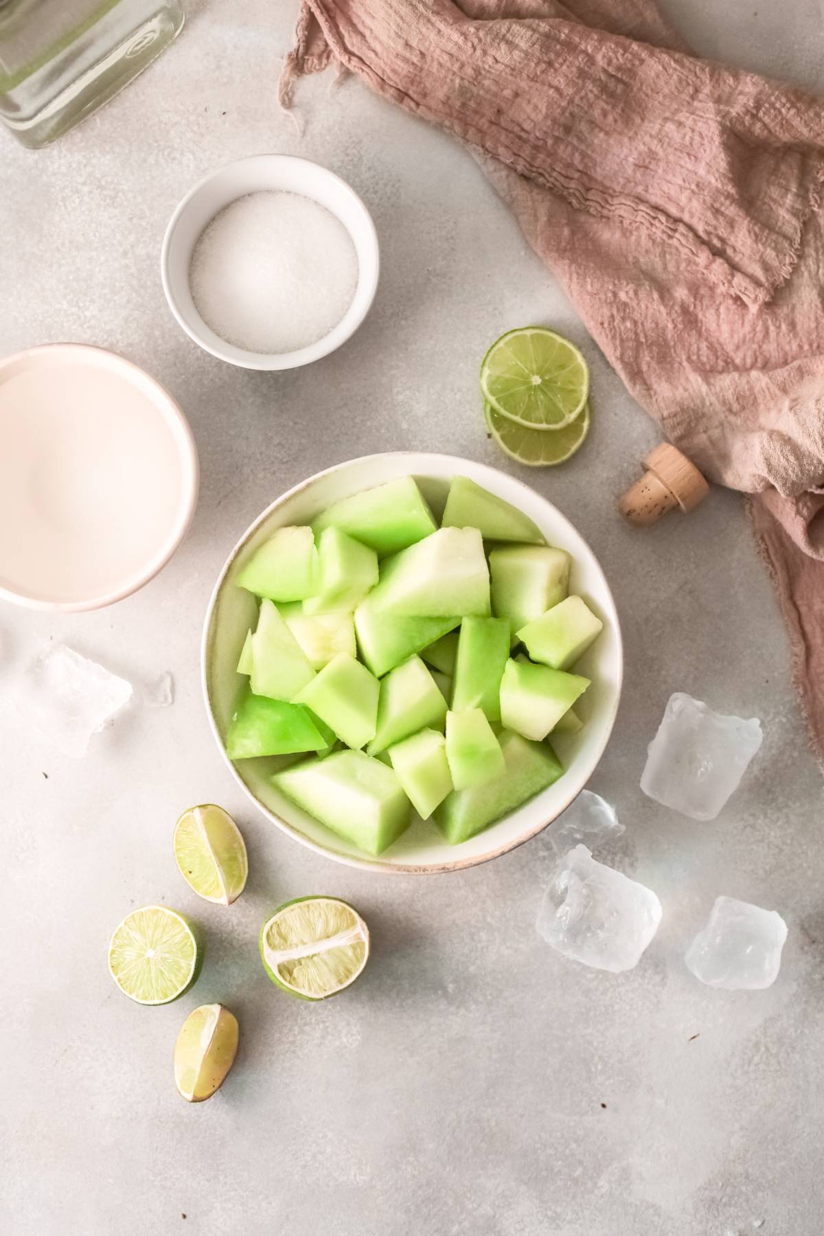 A bowl of chopped honeydew, lime halves, ice cubes, sugar, and a pink cloth on a light surface—perfect ingredients for a refreshing honeydew margarita or summer cocktail recipe with tequila.