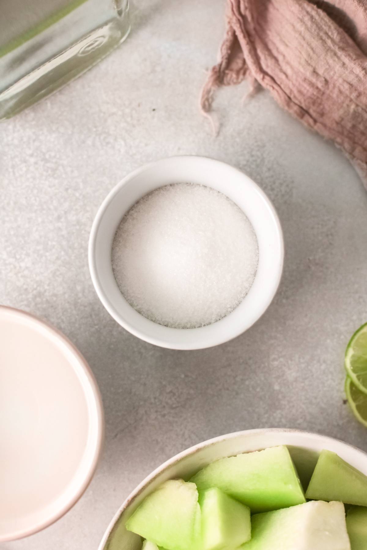 Small bowl of white sugar on a light surface, near honeydew pieces, a sliced lime, and a pink cloth—perfect ingredients for crafting a refreshing Honeydew margarita.