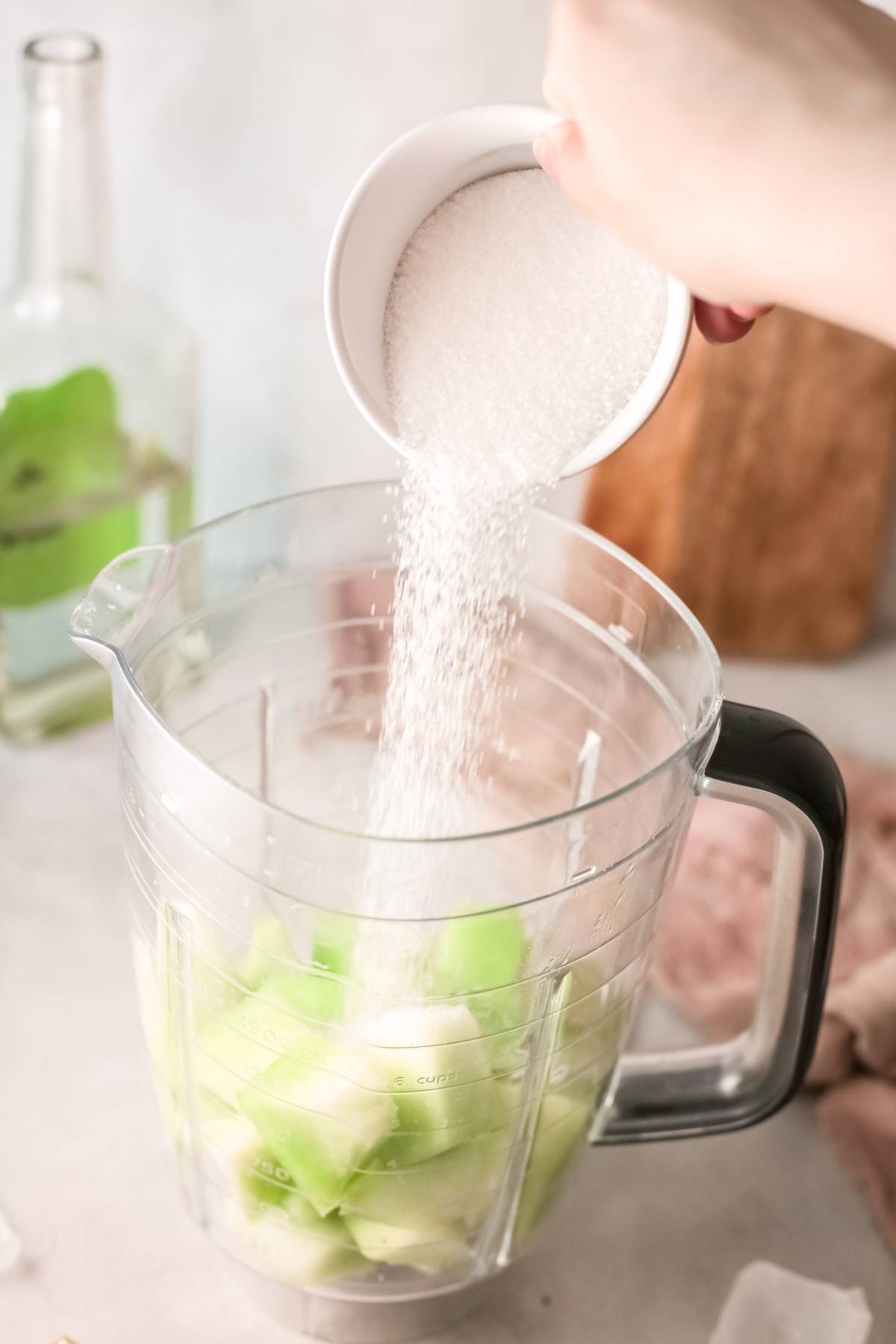 A hand pours sugar into a blender filled with chopped green fruit, likely honeydew melon, perfect for crafting an easy tequila cocktail or a refreshing summer cocktail recipe with tequila.