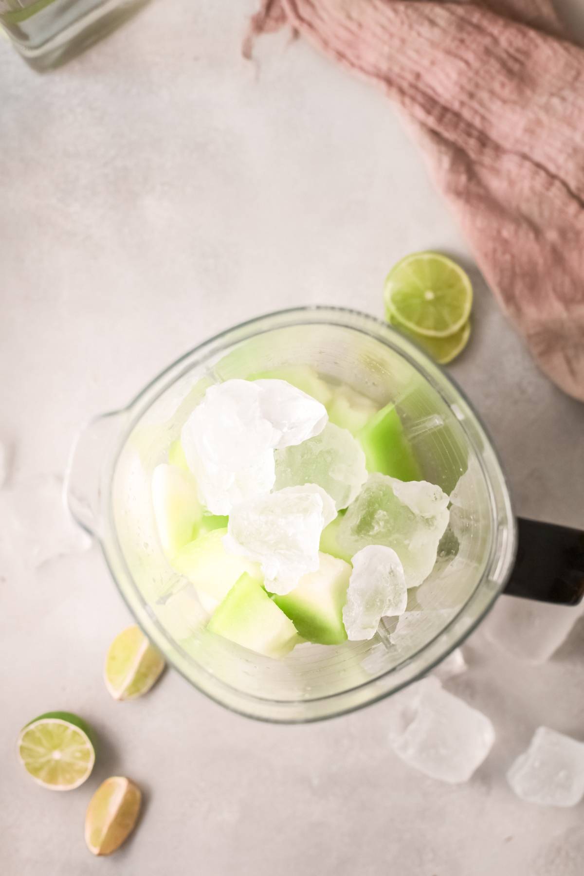 Top view of a blender with honeydew chunks, ice, and lime halves on a light countertop—perfect for blending into a refreshing honeydew margarita or your favorite summer cocktail recipe with tequila.