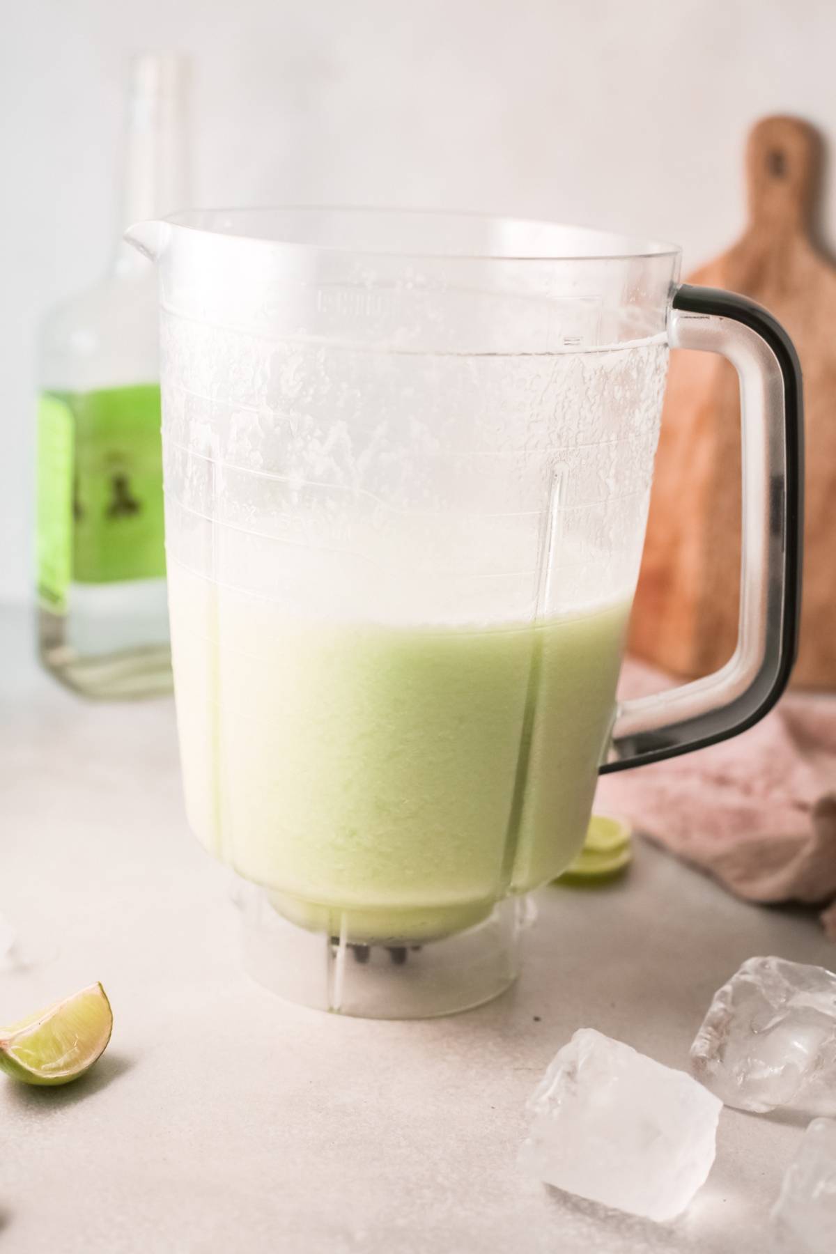 A blender pitcher filled with a light green honeydew margarita, surrounded by ice cubes and a lime wedge.