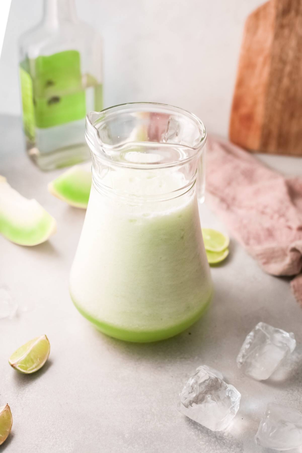 A glass pitcher of light green, frothy honeydew cocktail sits on a counter with lime wedges, ice cubes, and a bottle nearby—the perfect summer cocktail recipe with tequila.