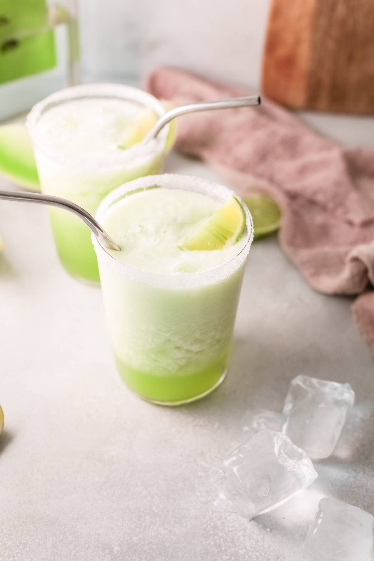 Two glasses of frothy honeydew cocktail with metal straws and lime wedges sit next to ice cubes on a light surface—an easy tequila cocktail perfect for summer.