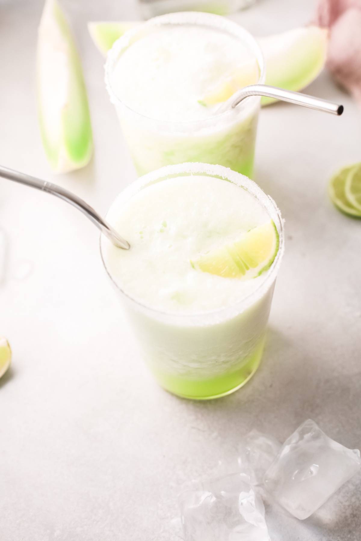 Two frosty green honeydew margaritas with lime wedges and metal straws sit on a light surface, surrounded by ice and lime slices—a refreshing summer cocktail recipe with tequila.