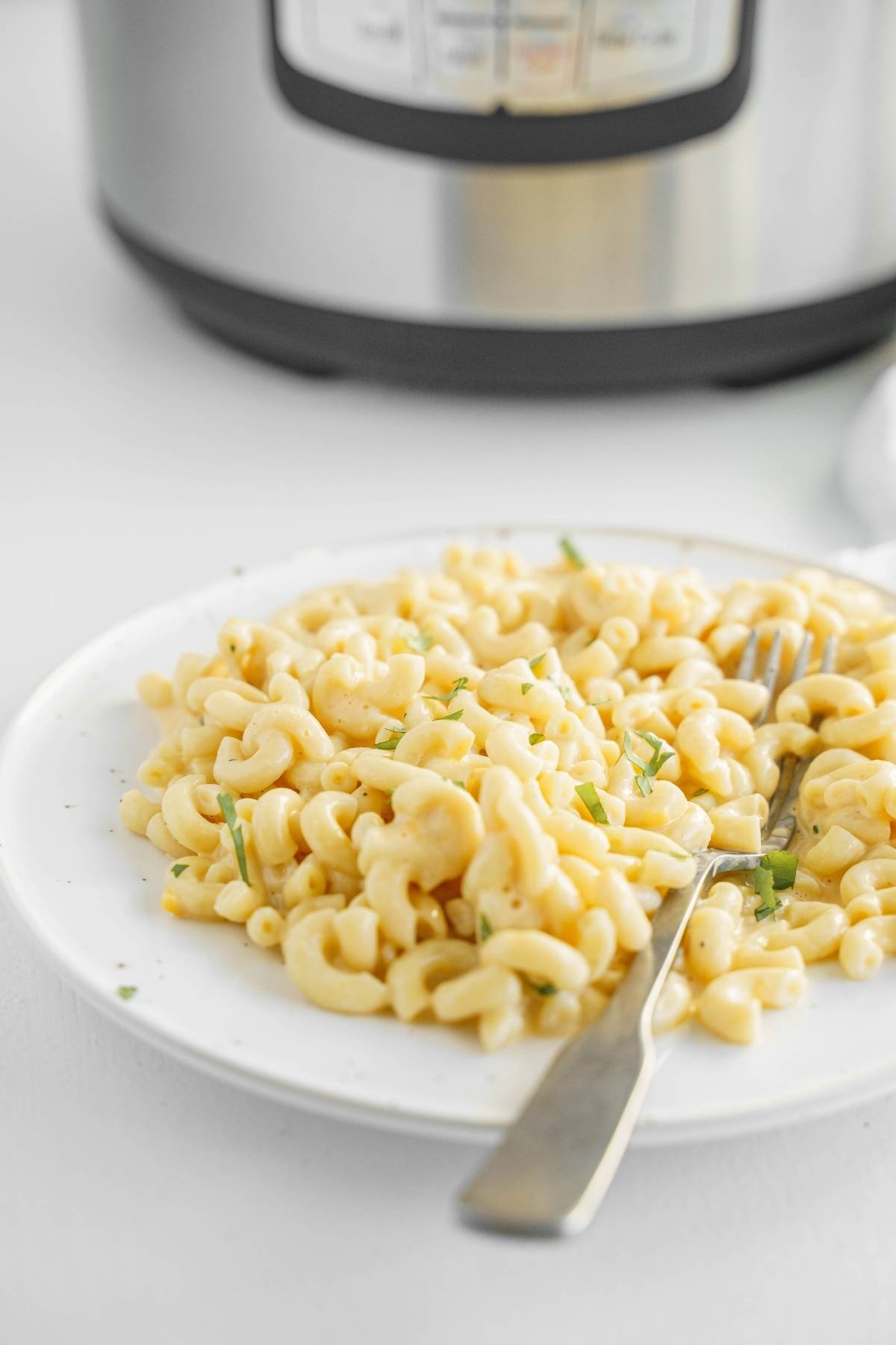 A plate of creamy Mac and cheese with herbs and a fork sits in front of a pressure cooker, perfect for anyone craving an easy instant pot pasta recipe.