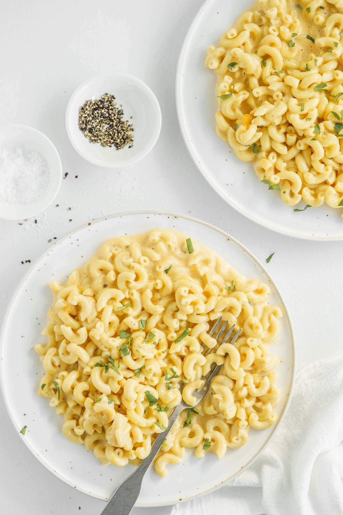 Two plates of mac and cheese garnished with herbs sit next to bowls of salt and pepper on a white table—perfect for anyone craving a classic or looking to try an Instant Pot macaroni and cheese recipe.