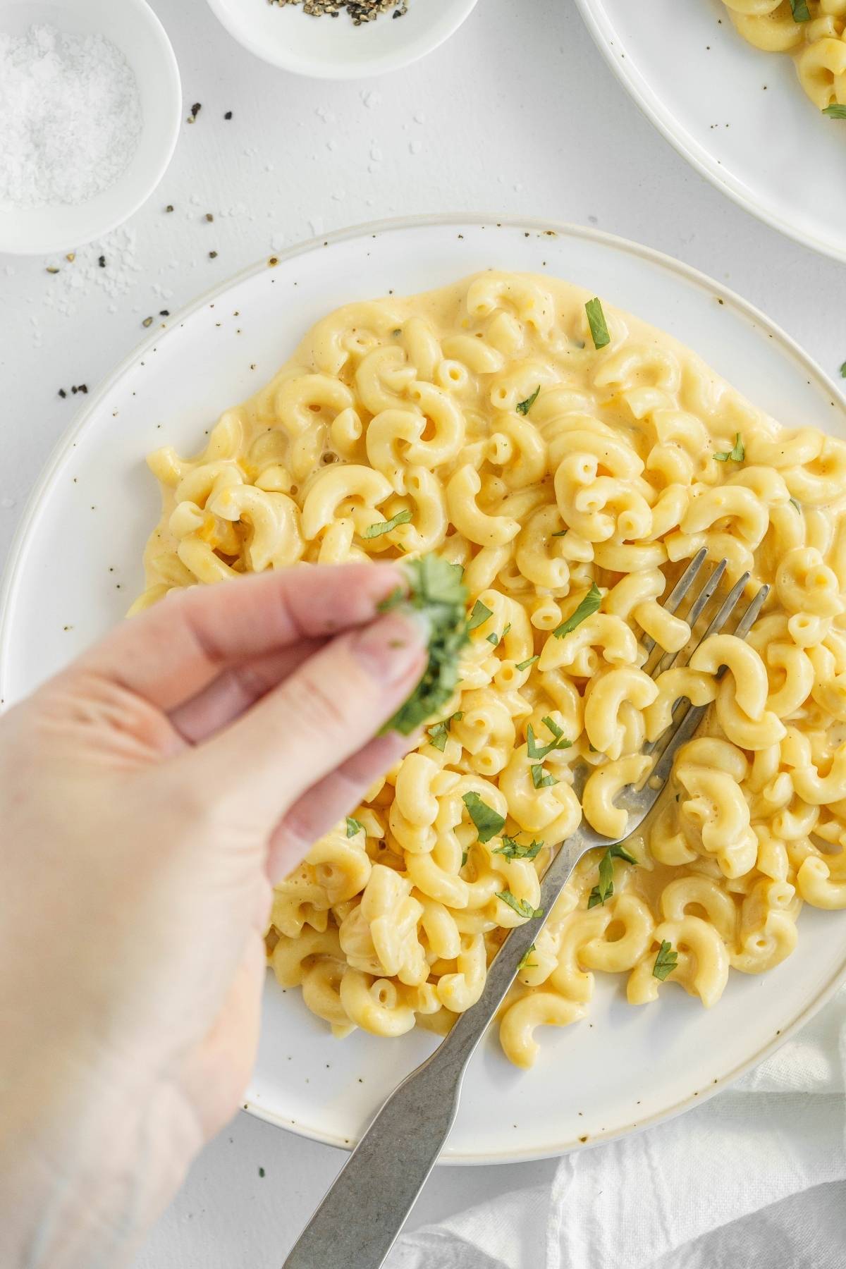 A hand sprinkles herbs over a plate of creamy Instant Pot macaroni and cheese, with a fork and knife on the side.