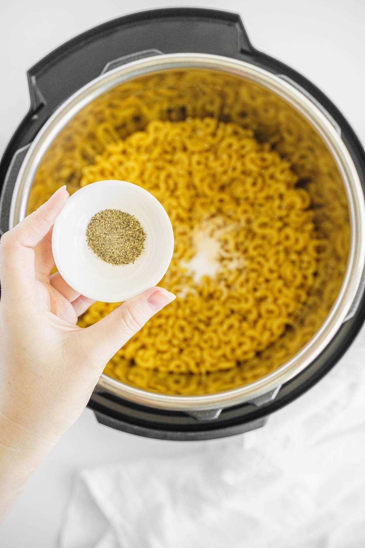 A hand holds a small bowl of pepper over uncooked macaroni in an Instant Pot, ready to begin an easy macaroni Instant Pot recipe.