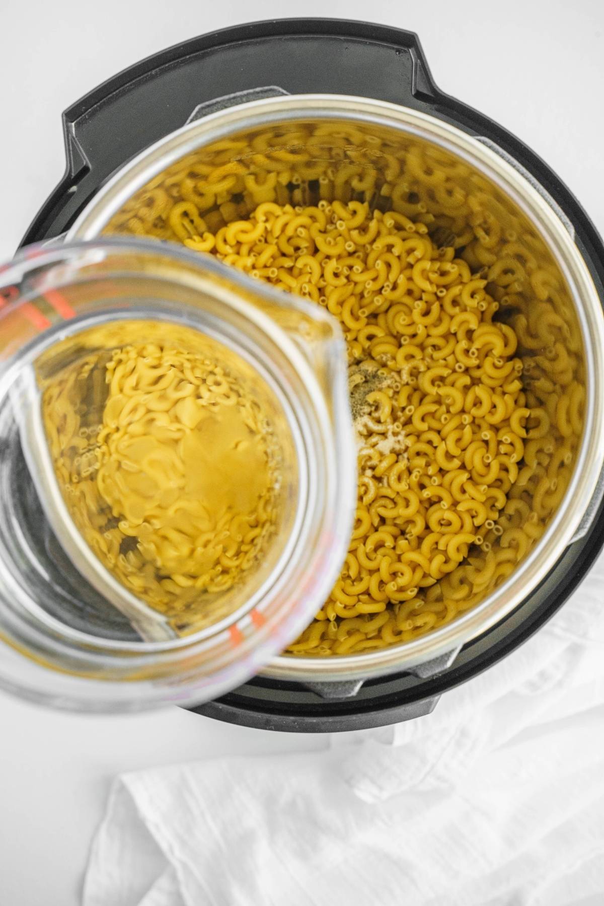 Overhead view of dry elbow macaroni in an Instant Pot as water is poured from a measuring cup—perfect for starting your favorite macaroni Instant Pot recipe or prepping easy Instant Pot macaroni and cheese.