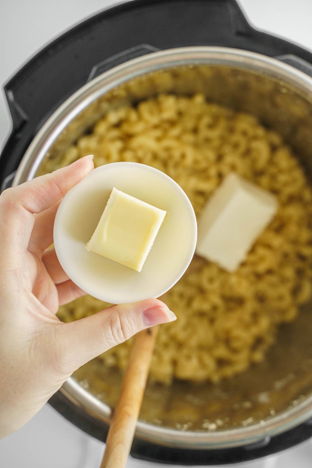 Hand holding a small bowl with a butter cube over a pot of cooked macaroni and another butter cube—perfect for making creamy Mac and cheese or your favorite easy Instant Pot pasta recipe.