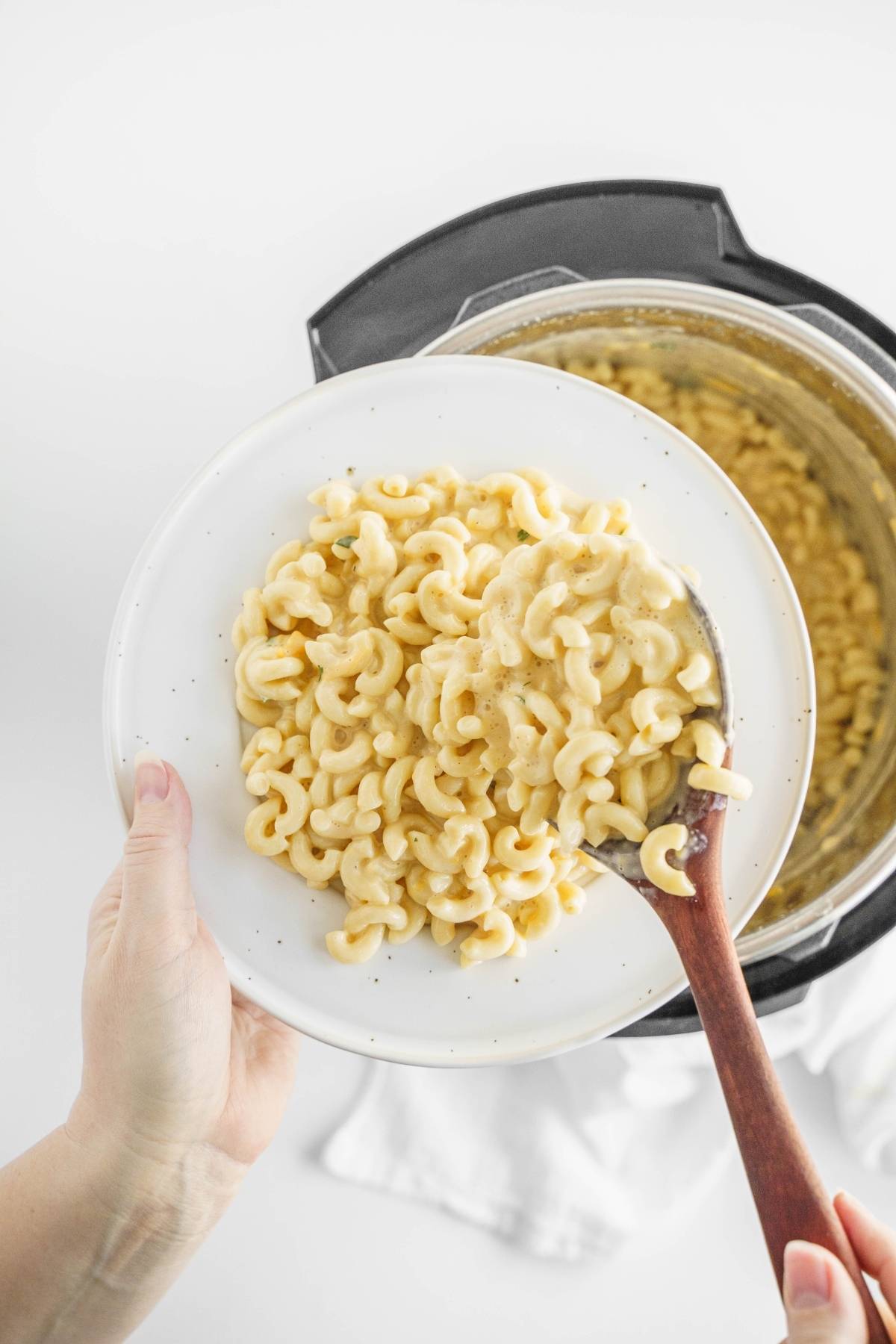 A hand serves creamy mac and cheese onto a white plate from an Instant Pot with a wooden spoon, showcasing an easy instant pot pasta recipe.