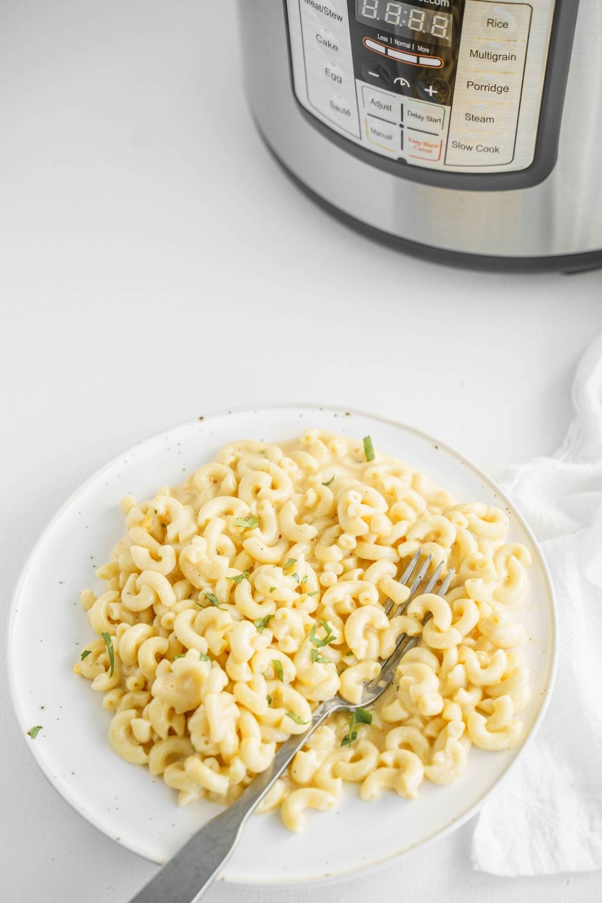 A plate of creamy mac and cheese with herbs and a fork sits beside an Instant Pot on a white surface, highlighting this easy instant pot pasta recipe.