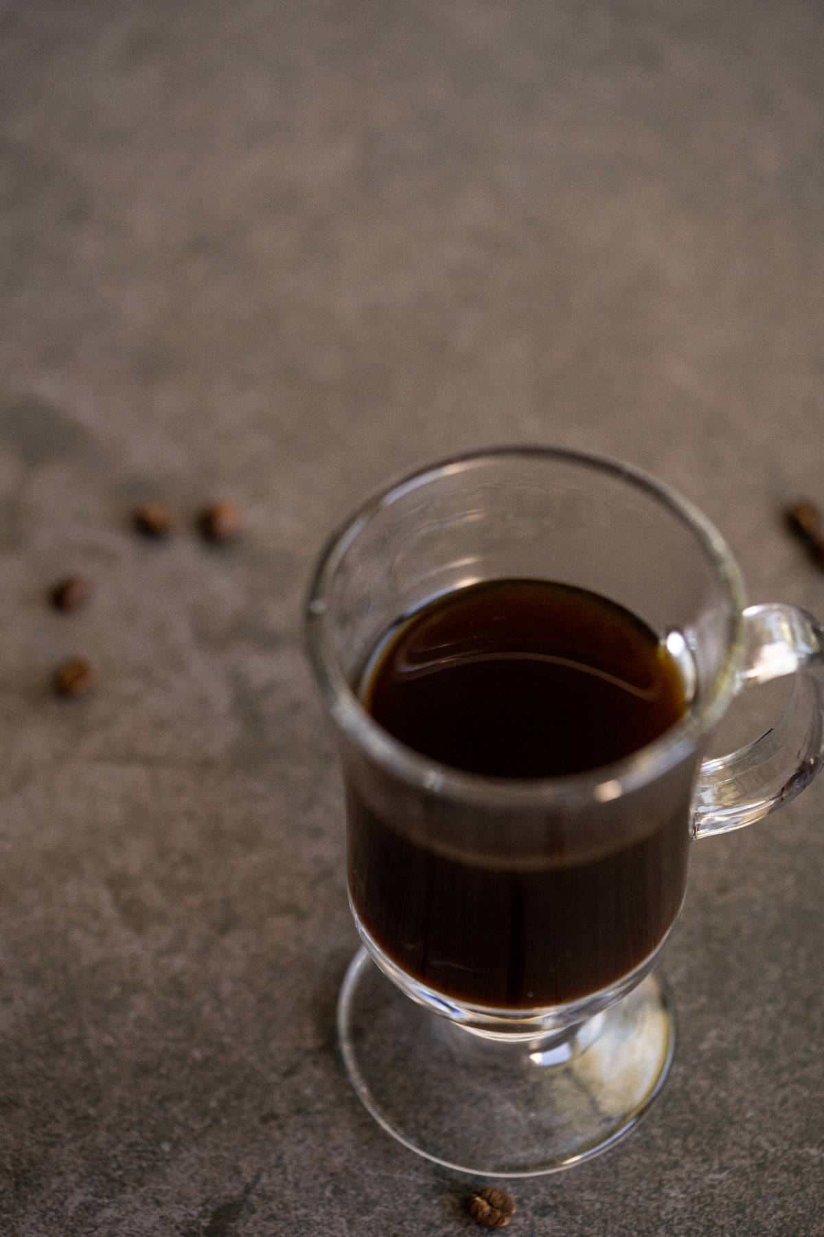 A glass mug of Irish cream coffee sits on a gray surface, with scattered coffee beans around it.