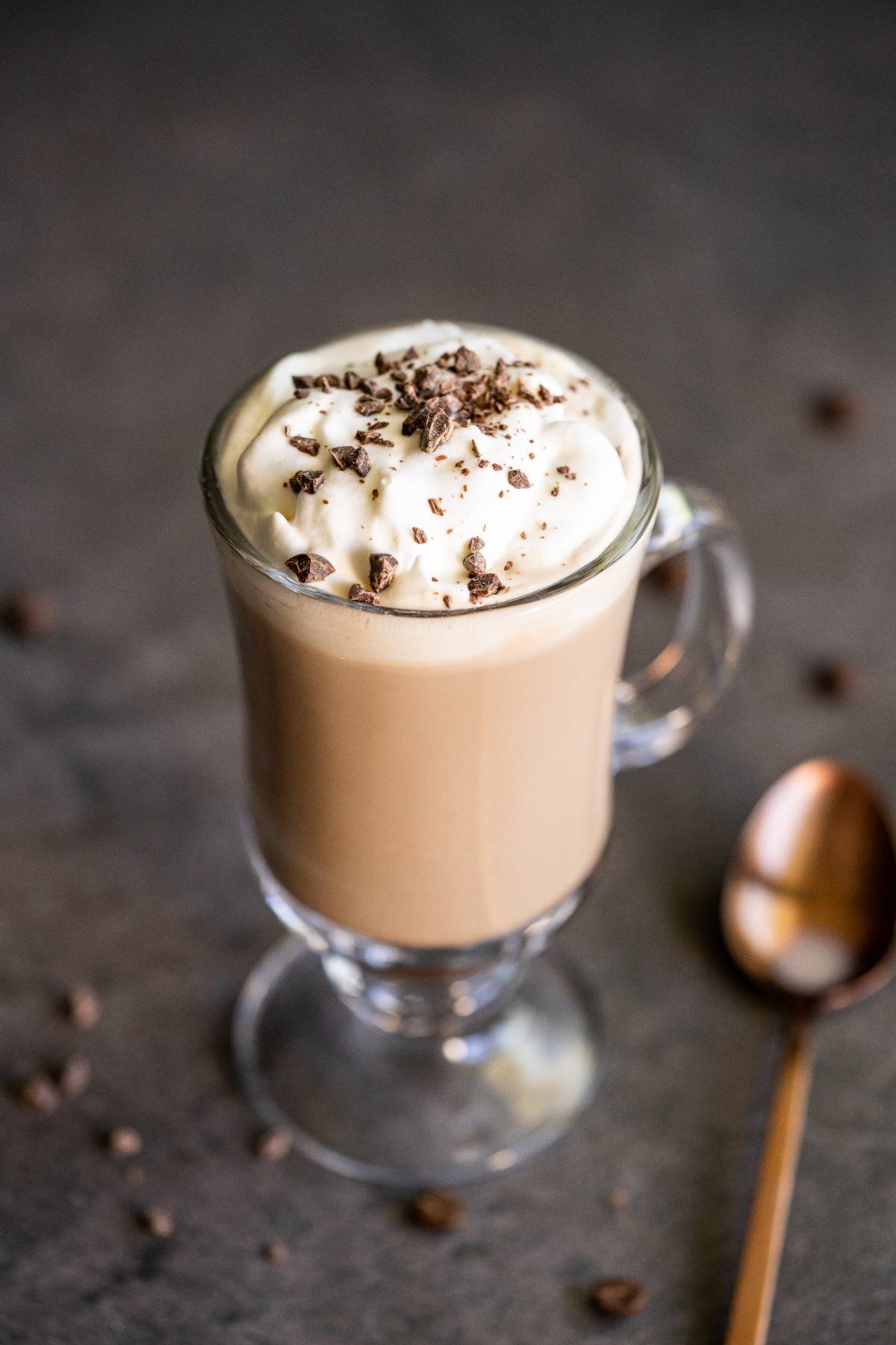Glass mug of creamy Bailey’s coffee recipe, topped with whipped cream and chocolate shavings, next to a spoon on a table.