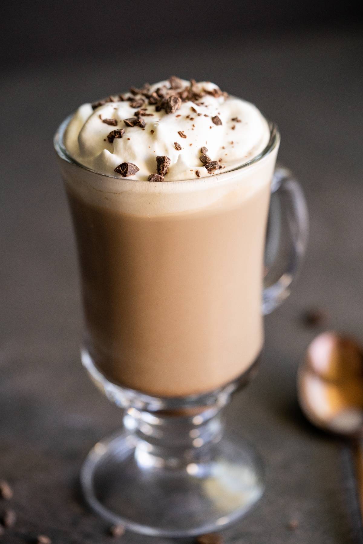 A glass mug of creamy Irish cream coffee topped with whipped cream and chocolate shavings, with a spoon nearby.