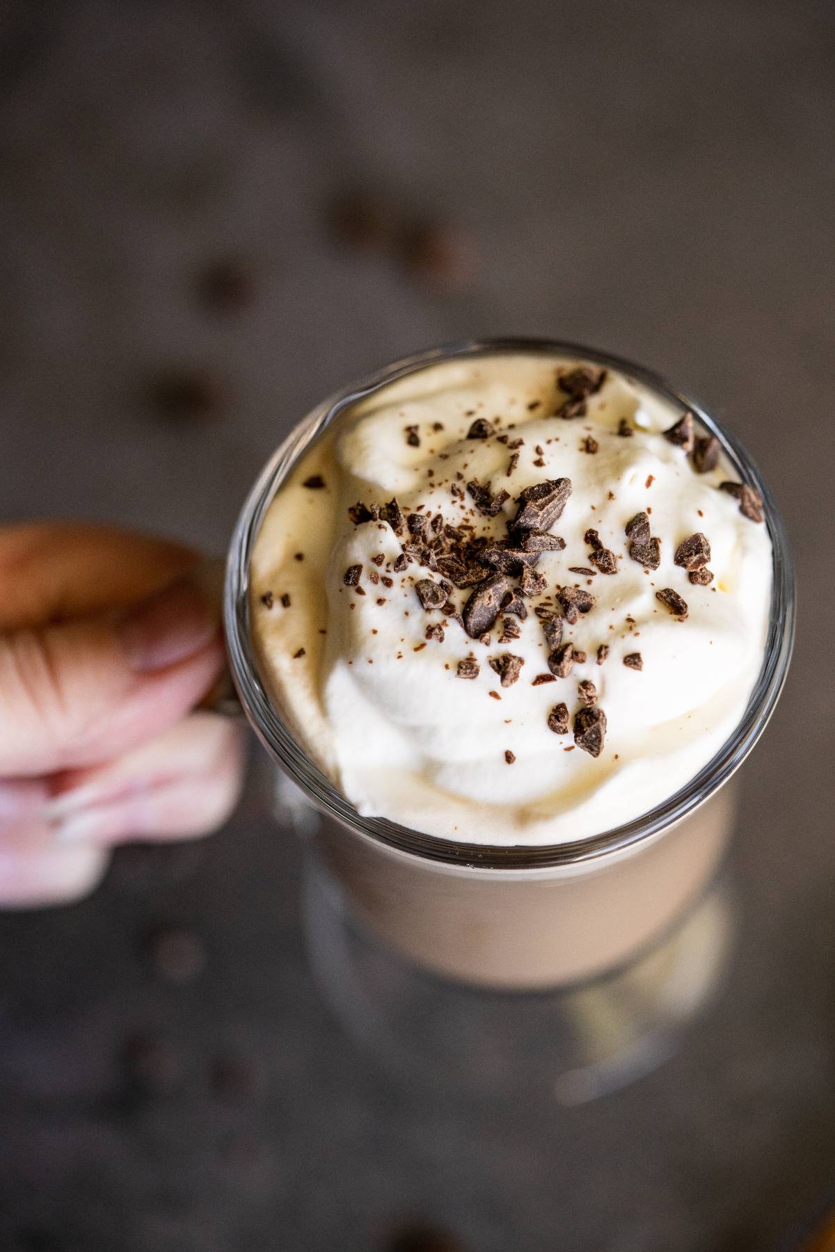 A hand holding a glass mug of hot chocolate topped with whipped cream and chocolate shavings, perfect for turning into a cozy Irish cream coffee or an indulgent alcoholic coffee drink.