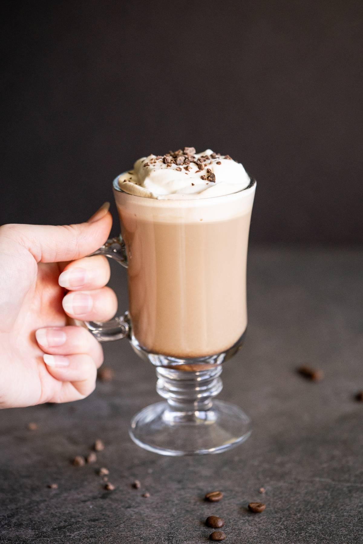 A hand holding a glass mug of Bailey’s coffee recipe—rich coffee topped with whipped cream and chocolate shavings—sits on a dark surface, perfect for those who love an indulgent alcoholic coffee drink.