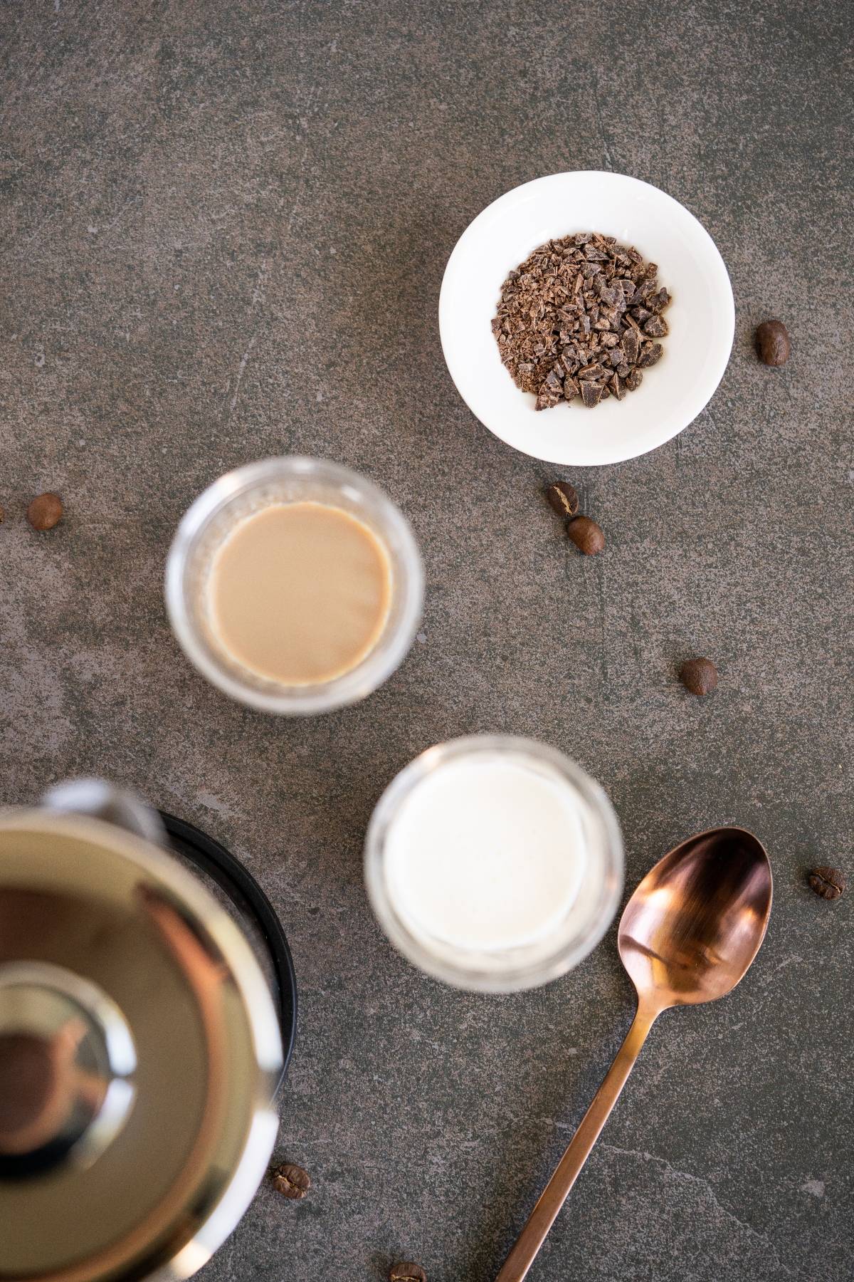 Two glasses with Irish cream coffee, a bowl of chocolate shavings, coffee beans, and a spoon rest on a gray surface.