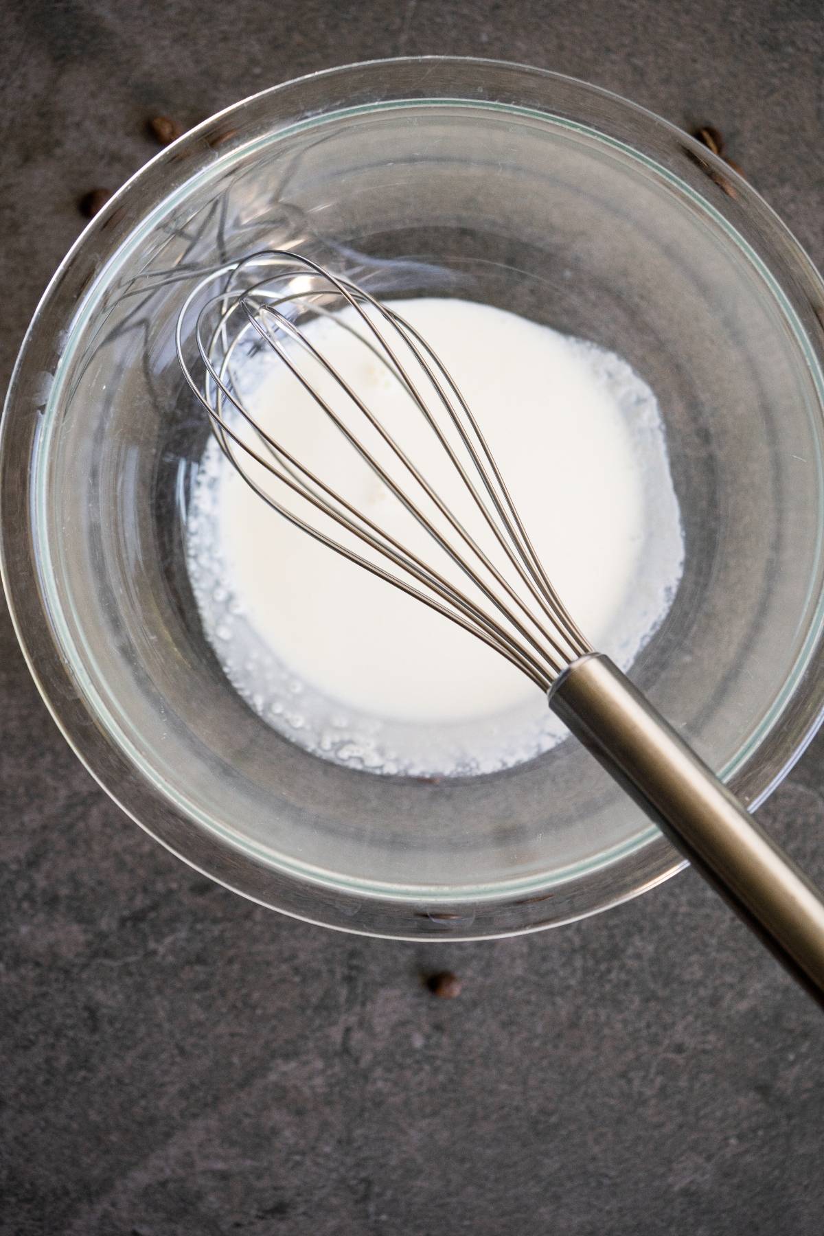 A glass bowl with milk and a metal whisk on a dark countertop, ready for creating the perfect Bailey’s coffee recipe.