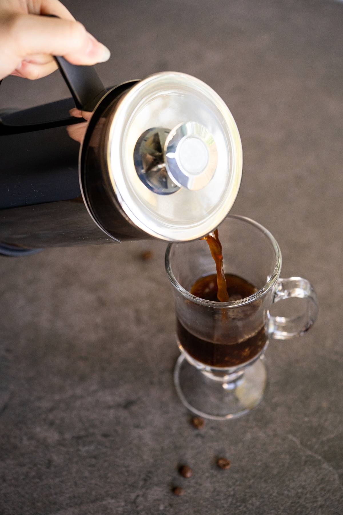 Coffee is being poured from a French press into a clear glass mug on a gray surface, perfect for enjoying an Irish cream coffee.