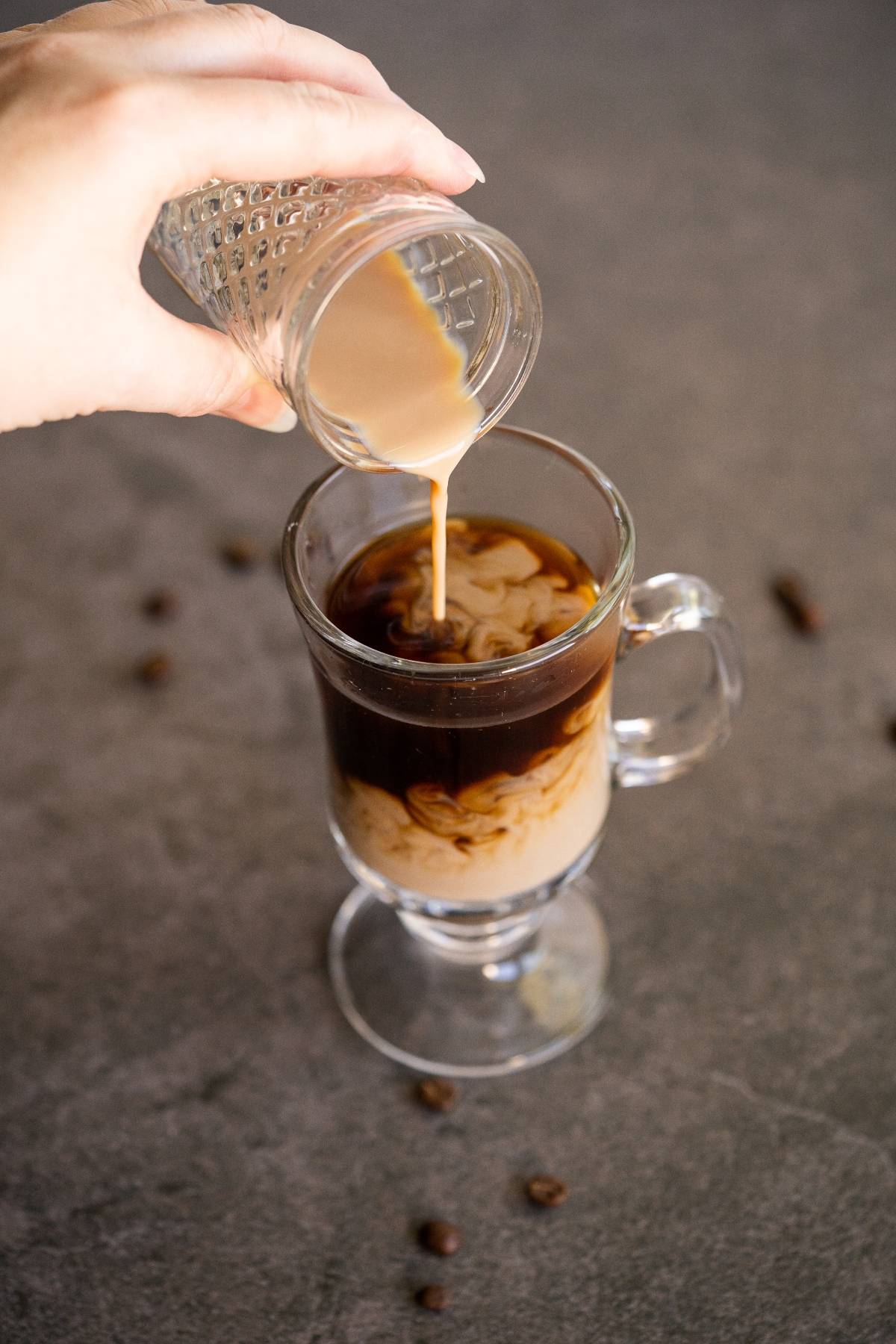 A hand pours cream into a glass mug of Irish cream coffee on a gray surface with scattered coffee beans.
