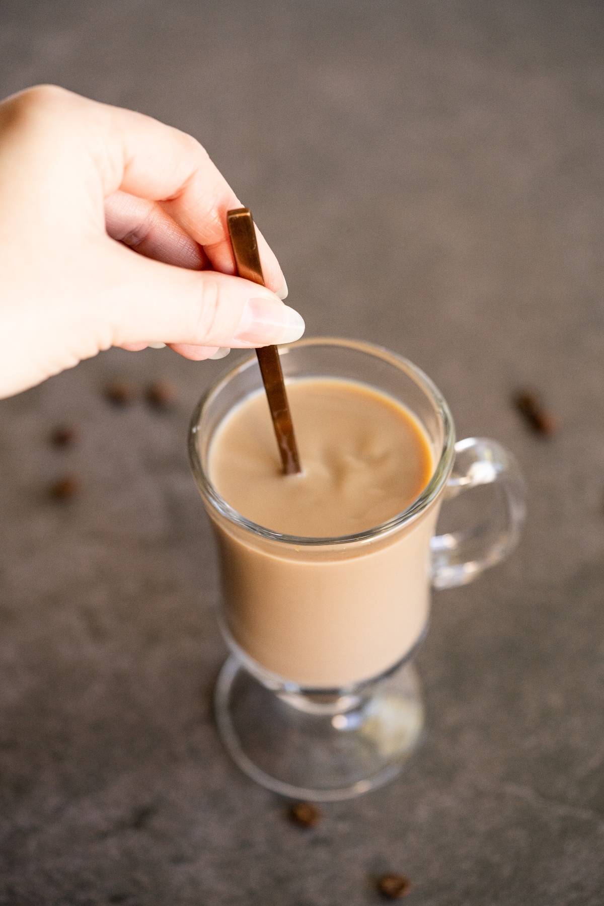 A hand stirs a glass mug of creamy Irish cream coffee with a metal spoon on a gray surface.