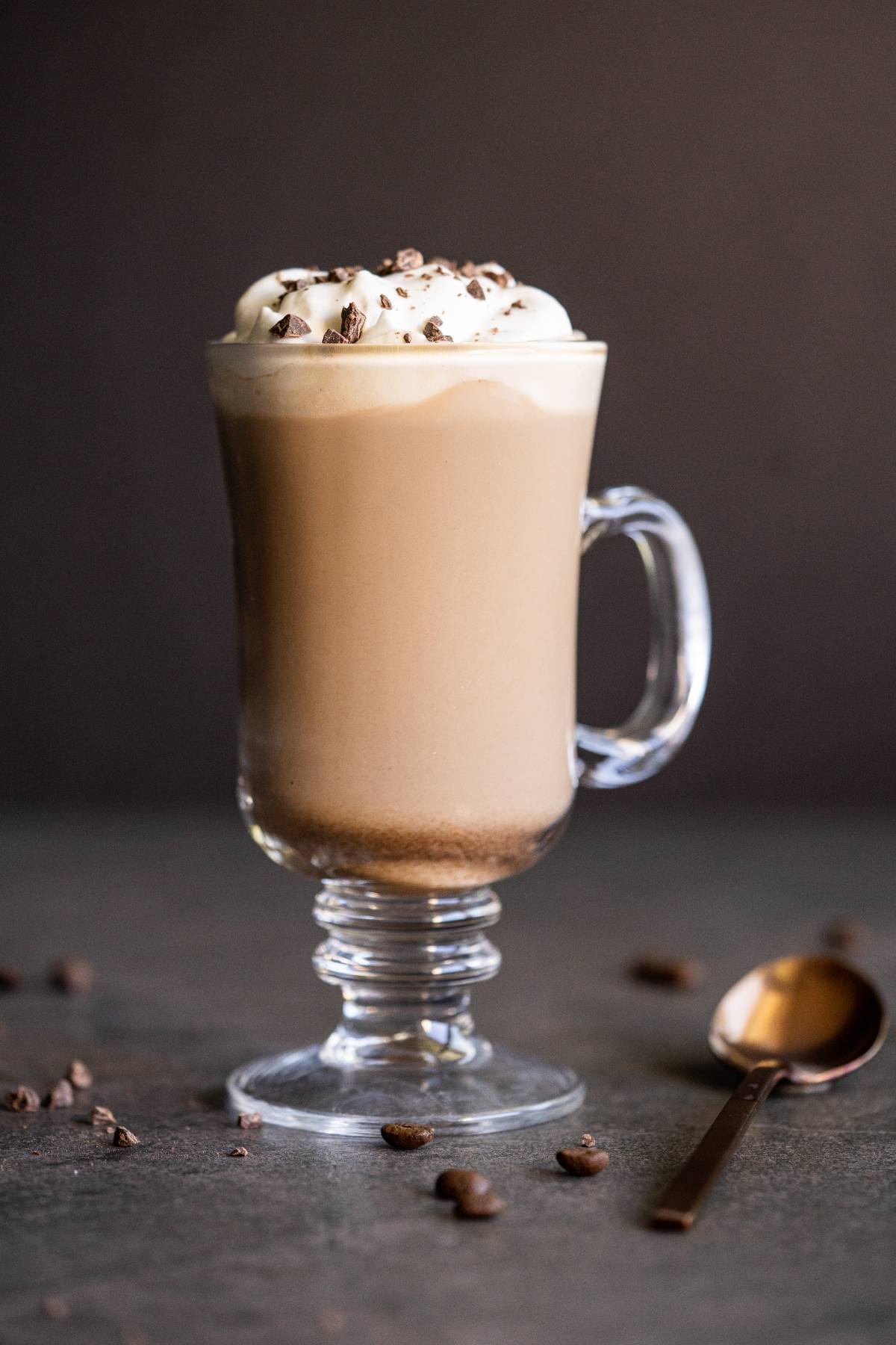 Irish coffee topped with whipped cream and chocolate shavings in a clear glass mug, this classic alcoholic coffee drink is served with coffee beans and a spoon nearby.