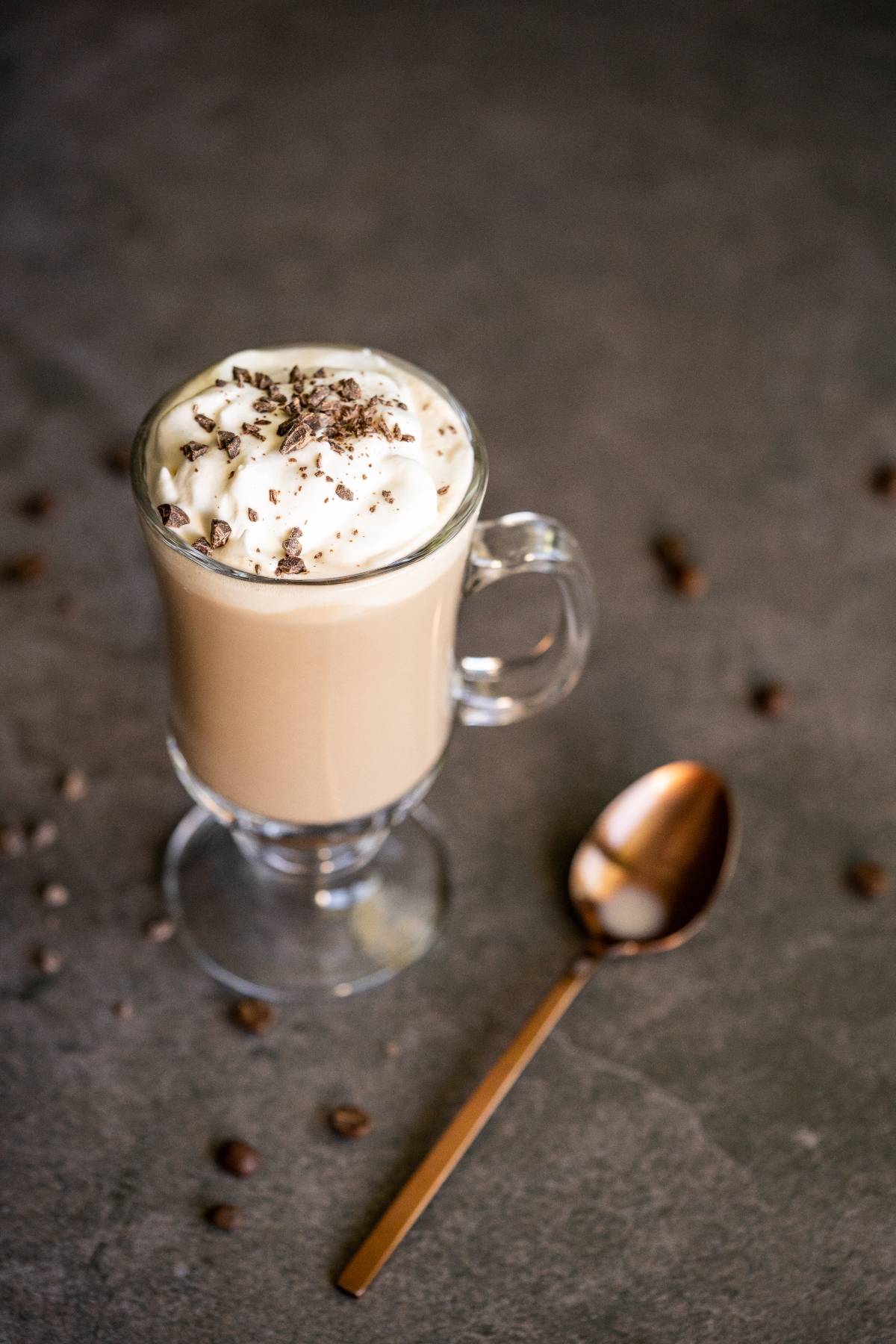 Glass mug of Irish cream coffee with whipped cream and chocolate shavings, next to a gold spoon on a dark surface.