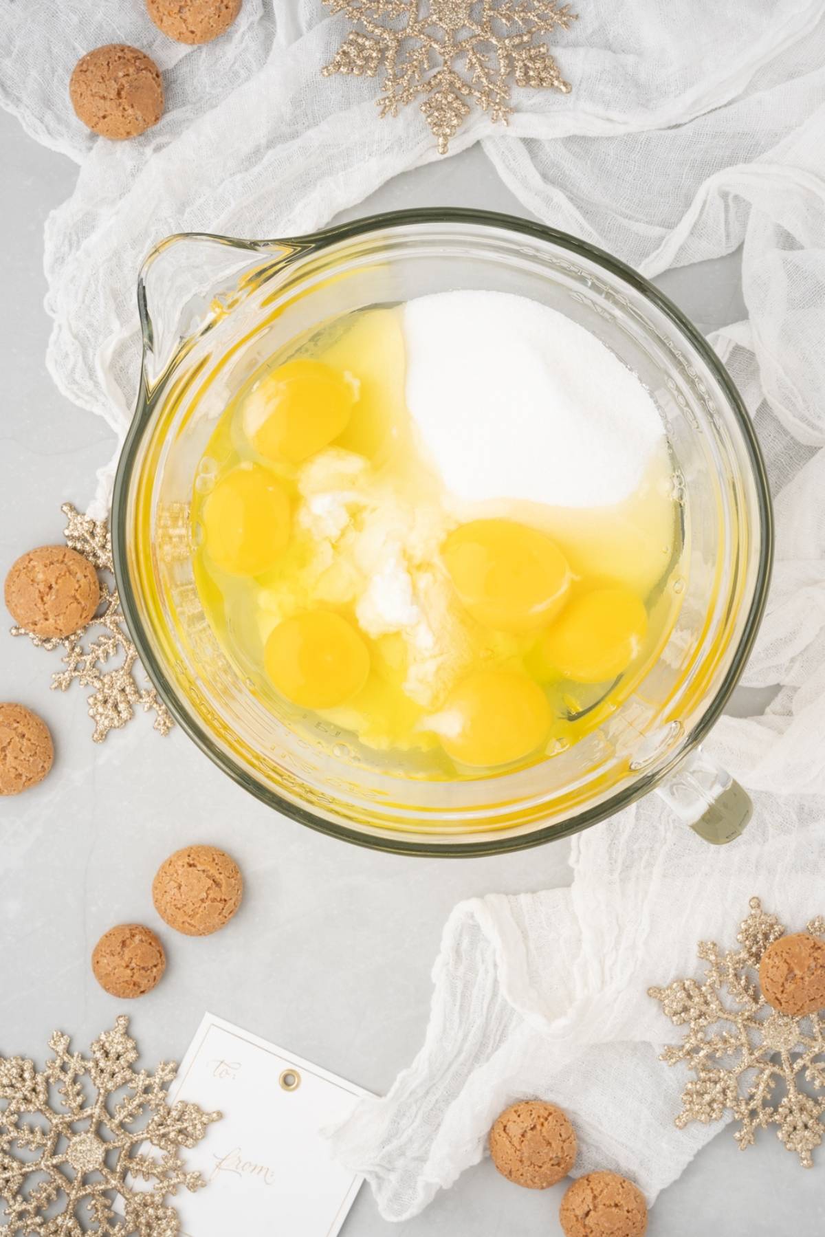 A glass bowl with eggs and sugar, surrounded by cookies, golden snowflakes, and white fabric—perfect for creating a classic Italian ricotta dessert made with amaretti cookies.