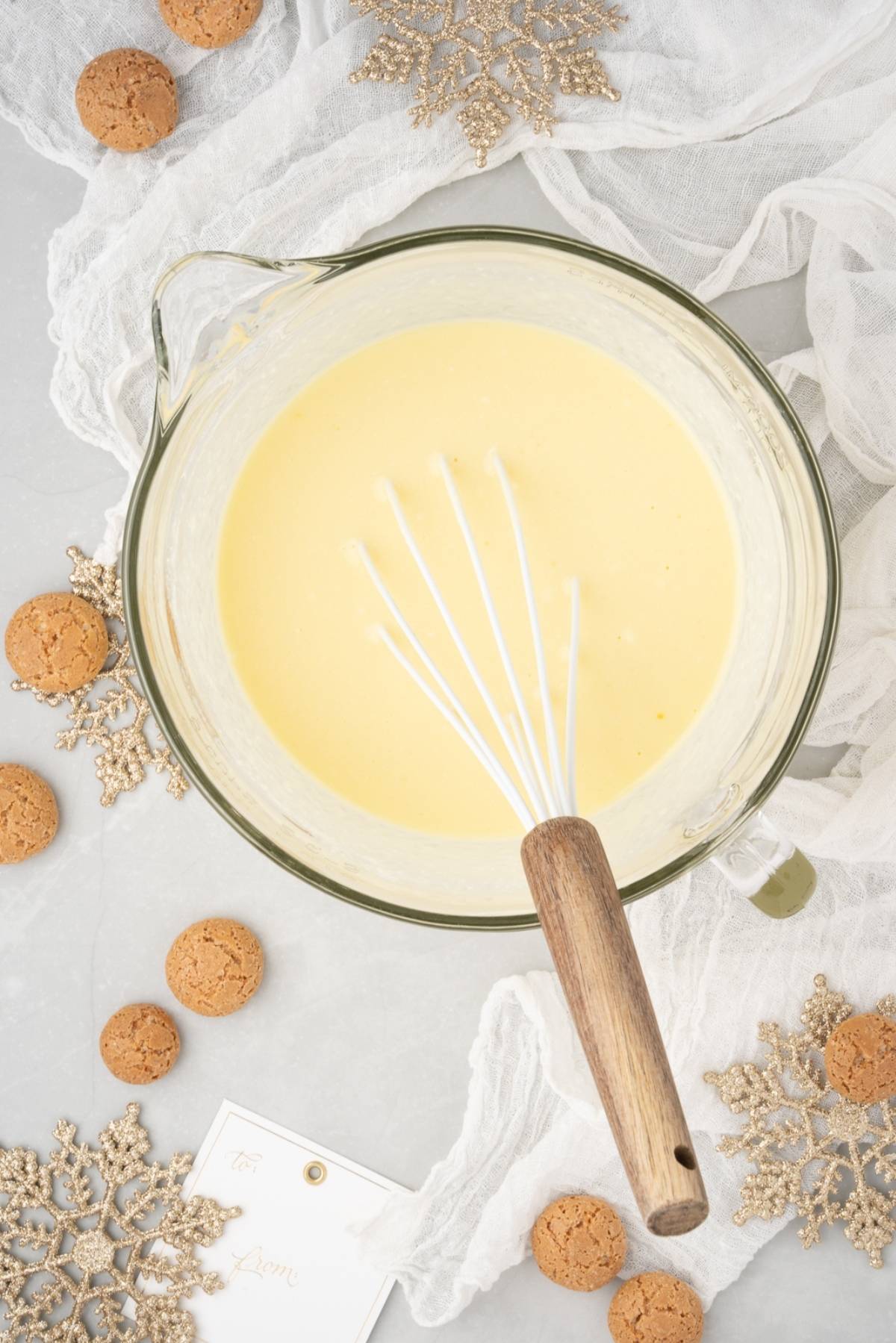 A glass bowl of yellow batter for Lemon ricotta amaretti cake with a whisk, surrounded by cookies and gold snowflake decorations, ready to be served at Christmas or other holidays.
