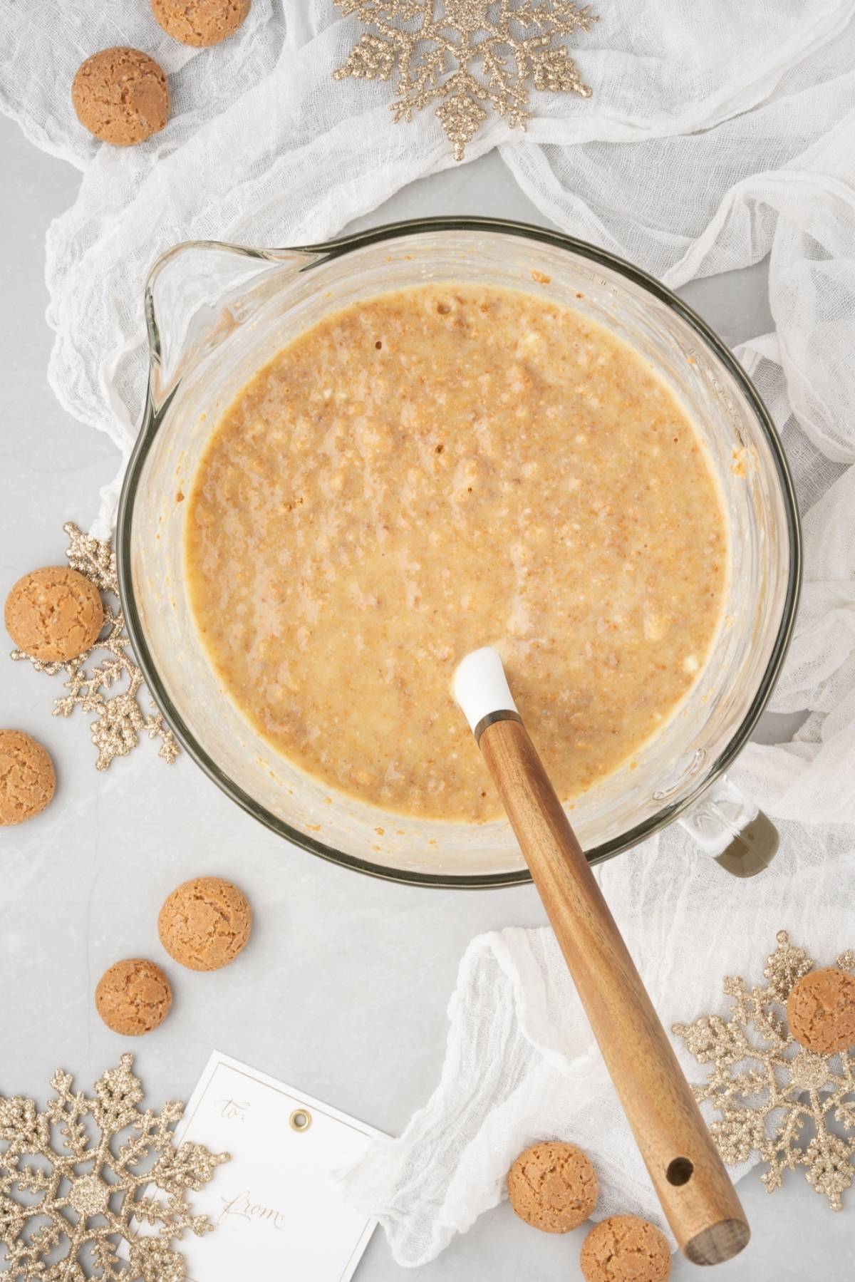 A glass bowl of cookie batter with a wooden spoon, surrounded by cookies and gold snowflake ornaments—perfect for making Lemon ricotta amaretti cake or other treats made with amaretti cookies and ricotta cheese.