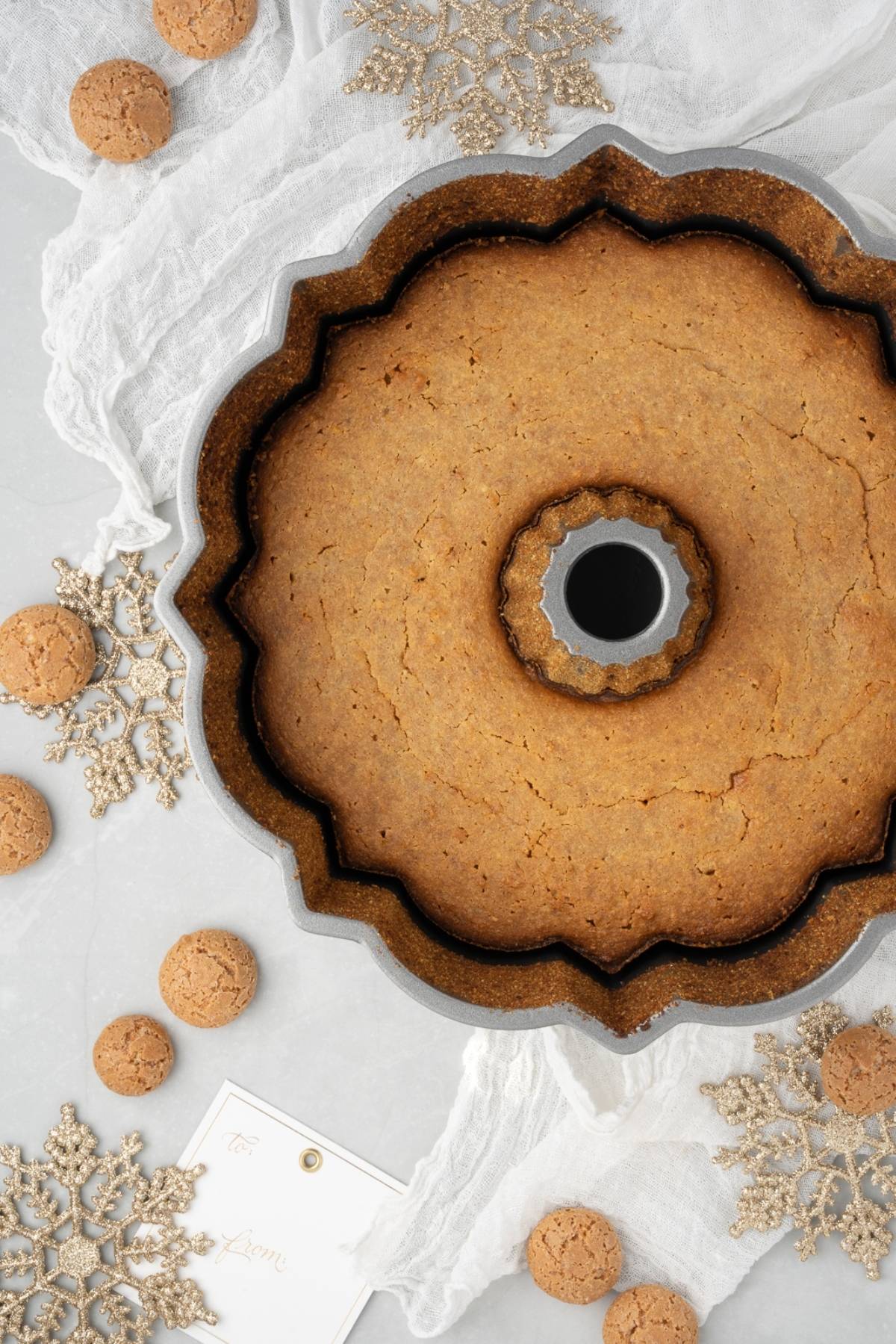 Top view of a Lemon ricotta amaretti cake in a pan, surrounded by cookies, snowflake ornaments, and a gift tag.