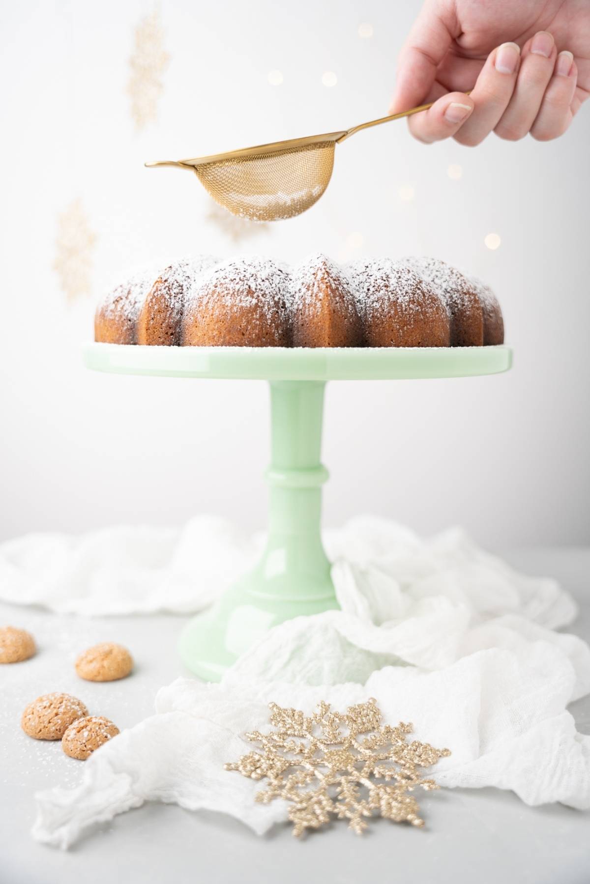 A hand sifts powdered sugar onto a Lemon ricotta amaretti cake on a pale green stand, with cookies and gold decor nearby.