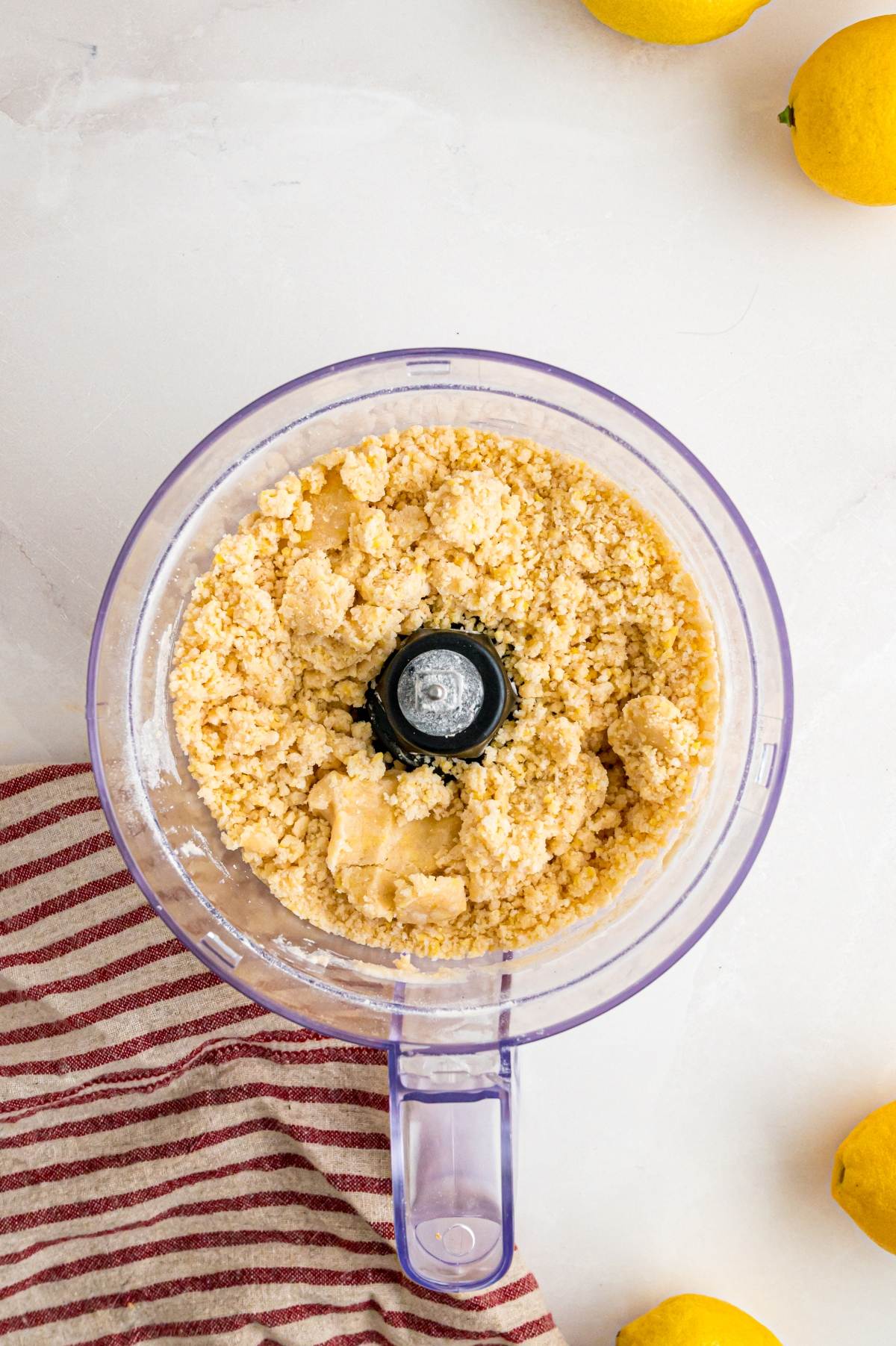 Food processor with crumbly dough, on a white surface near lemons and a red-striped kitchen towel.