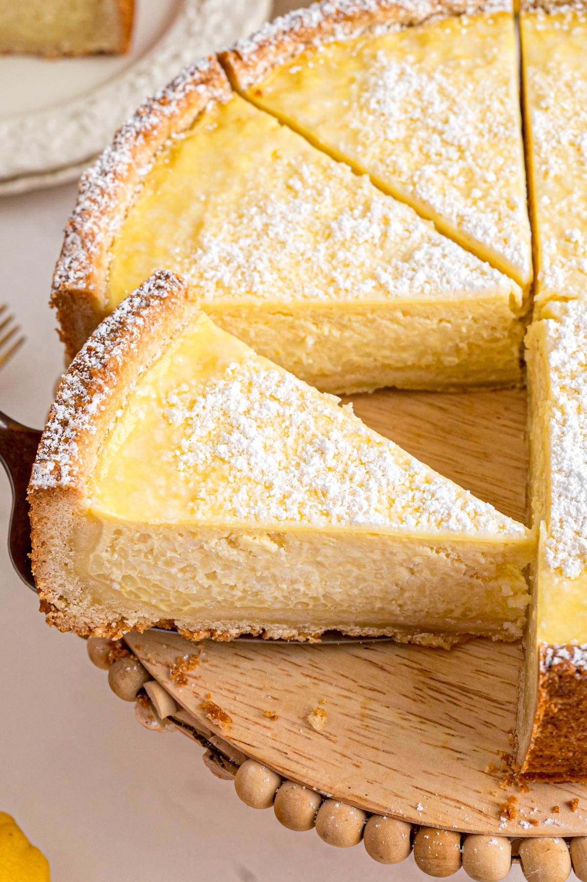 A slice of creamy cheesecake topped with powdered sugar being lifted from a wooden cake stand.