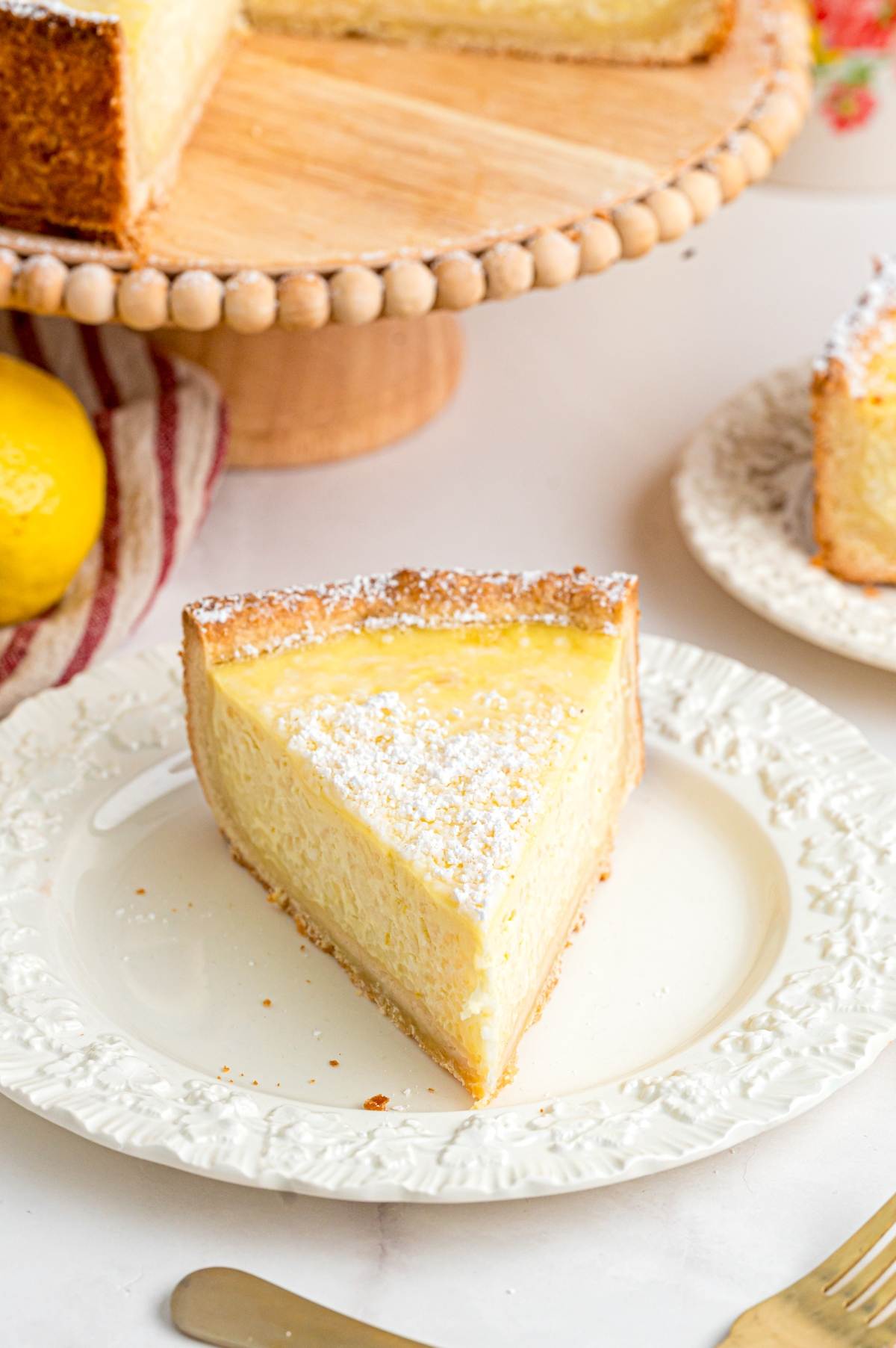 A slice of lemon cheesecake dusted with powdered sugar on a white plate, with more cake in the background.