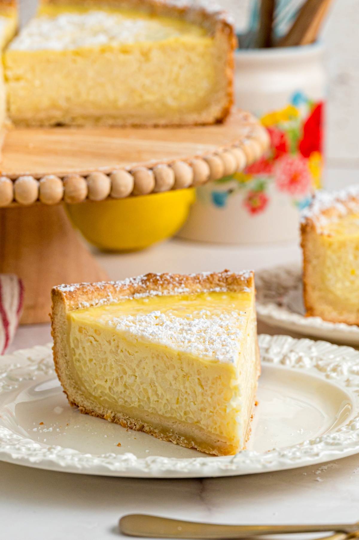 A slice of ricotta cheesecake with powdered sugar on a plate, in front of a cake stand with more cheesecake.